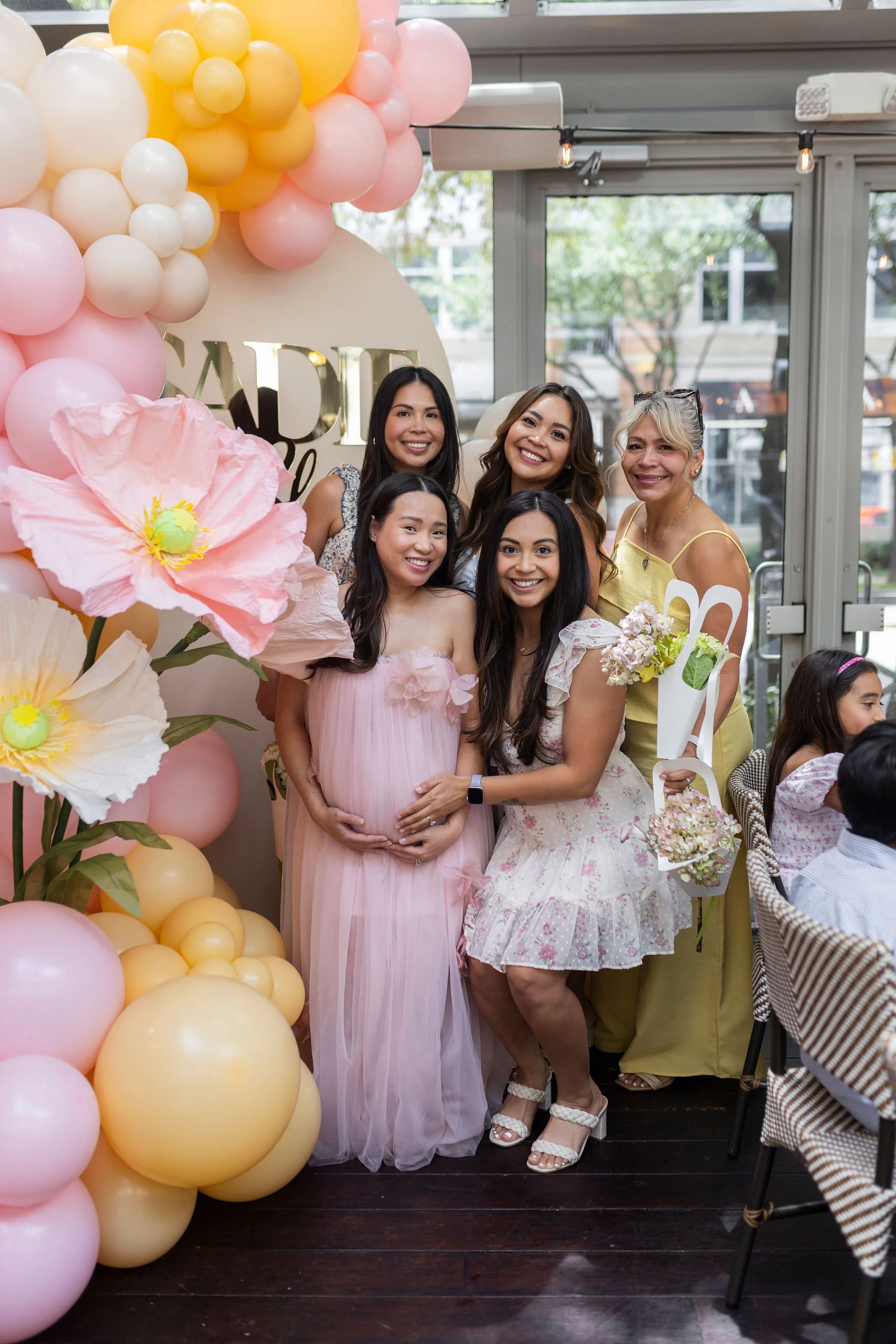 A group of five women celebrating a baby shower behind a pink and yellow balloon arch, with one woman holding her pregnant belly and another holding a bouquet of flowers.