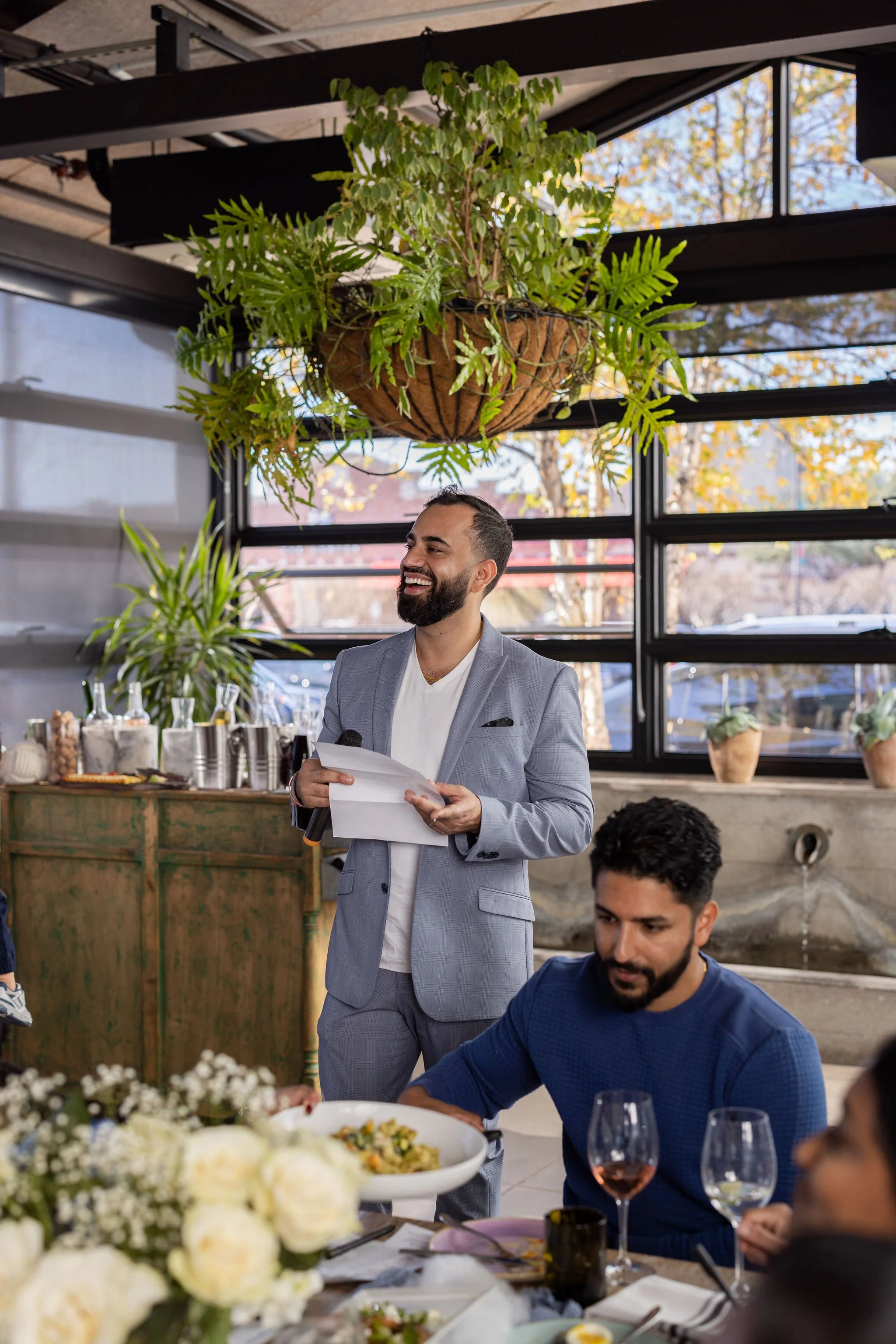 A man in a light gray suit standing and smiling while holding a piece of paper at a social gathering or banquet inside a modern venue with large windows and hanging plants. Other people are seated at the dining table with plates of food and glasses of wine.