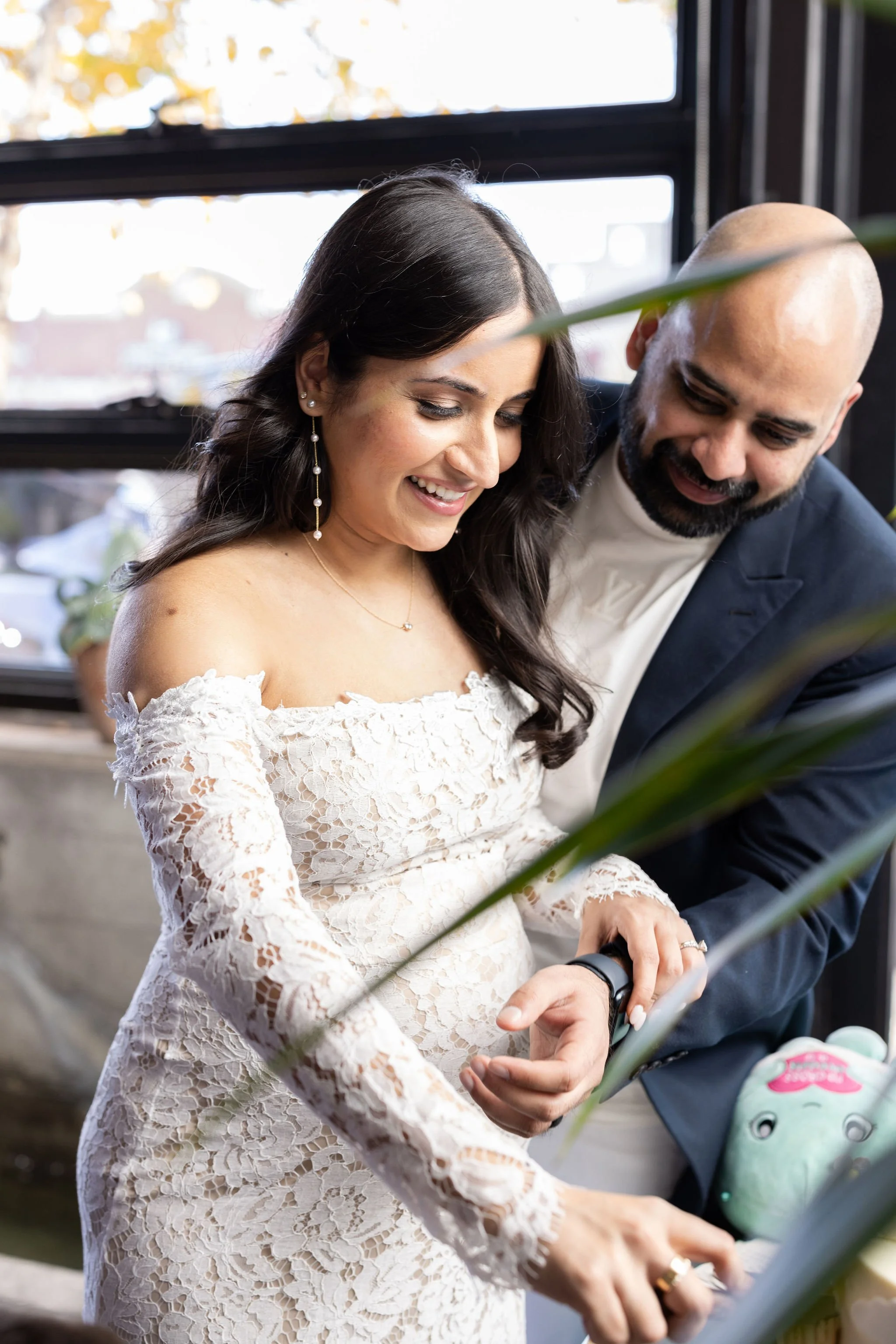 A bride and groom exchanging rings during their wedding ceremony, indoors near a window with natural light, with a plant partially visible in the foreground.