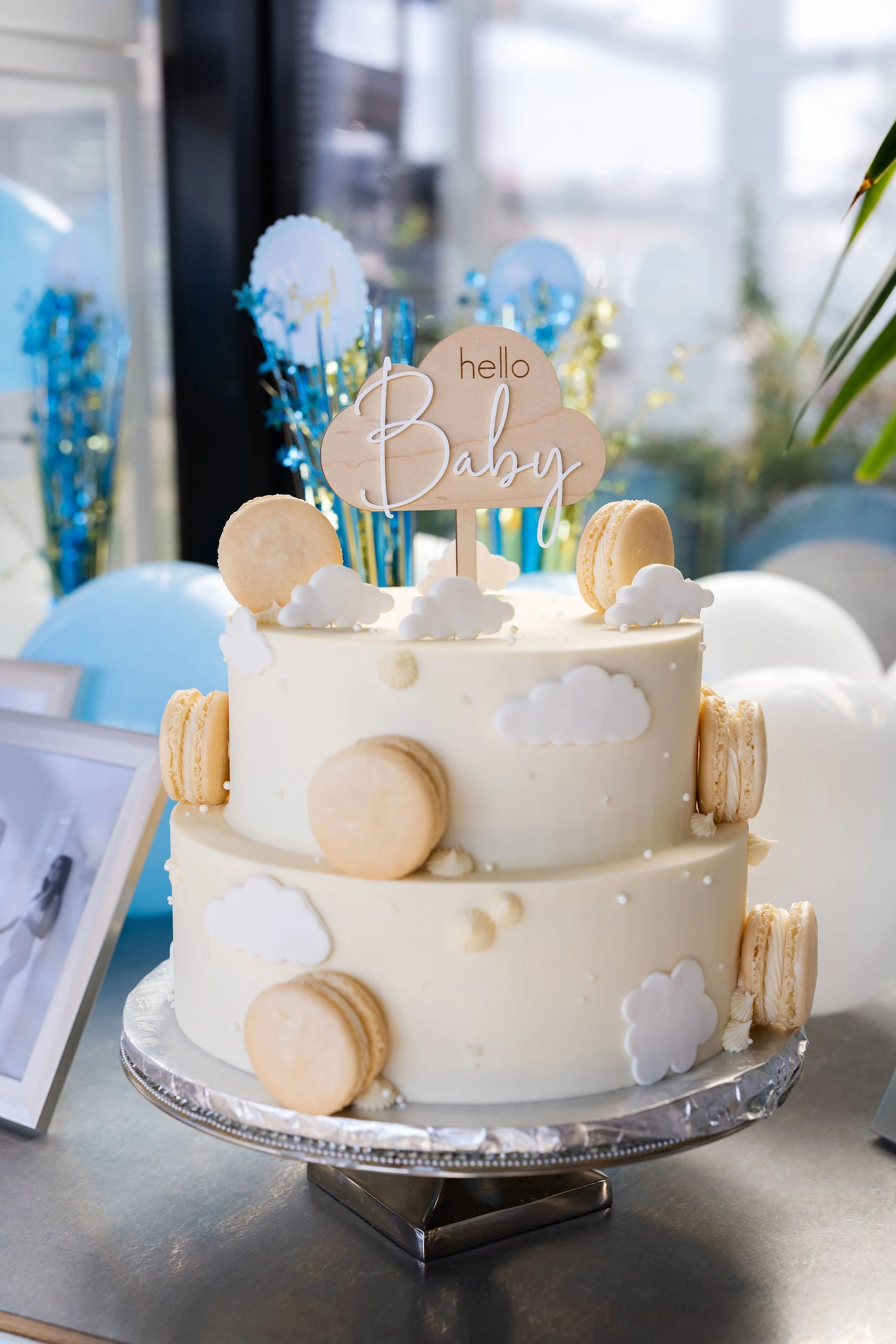 A white tiered cake decorated with small cloud-shaped fondant and macarons, with a wood sign reading 'hello Baby' in cursive, set on a silver cake stand.