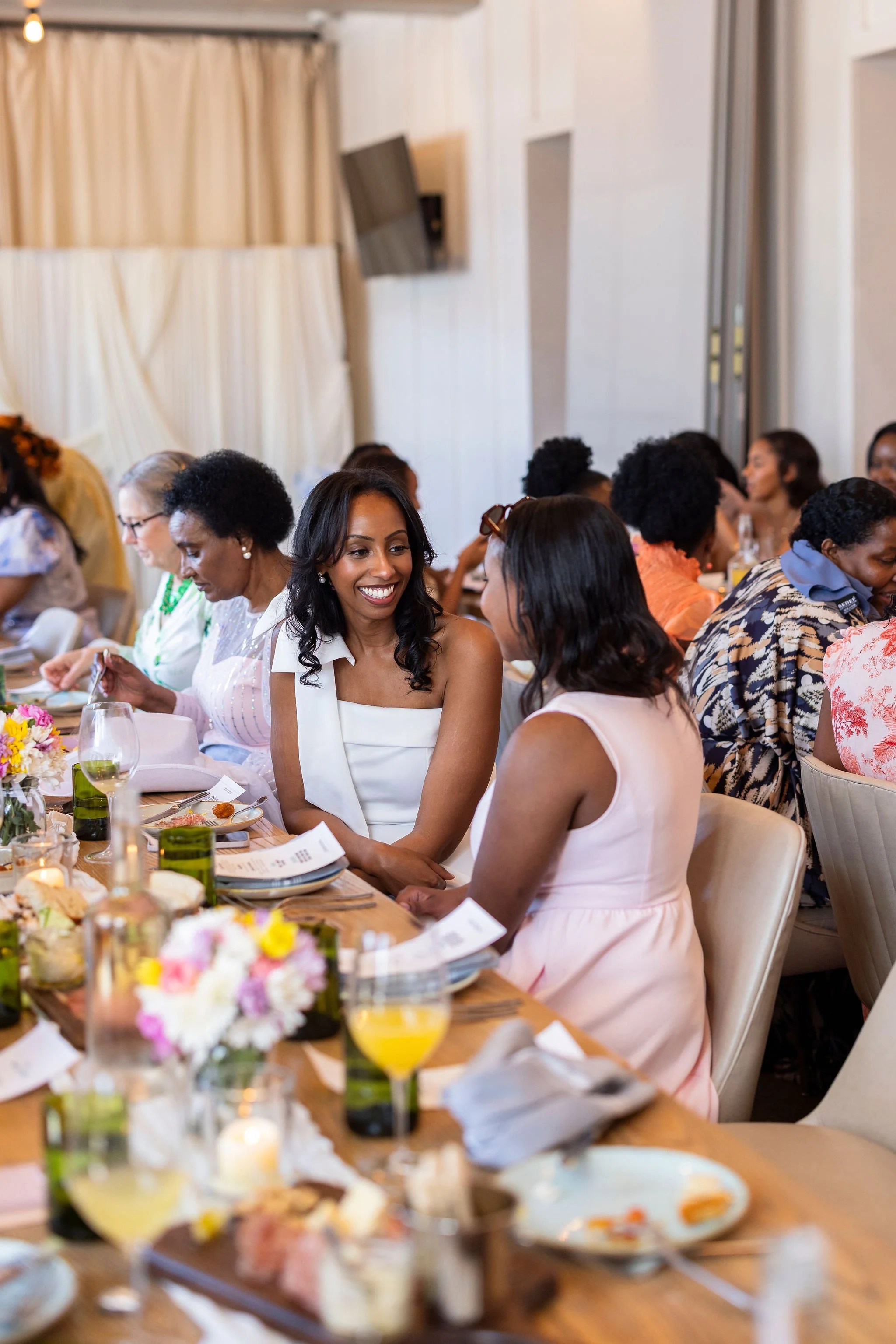 Women sitting around a dining table at a social gathering or celebration, smiling and talking, with food and drinks on the table.
