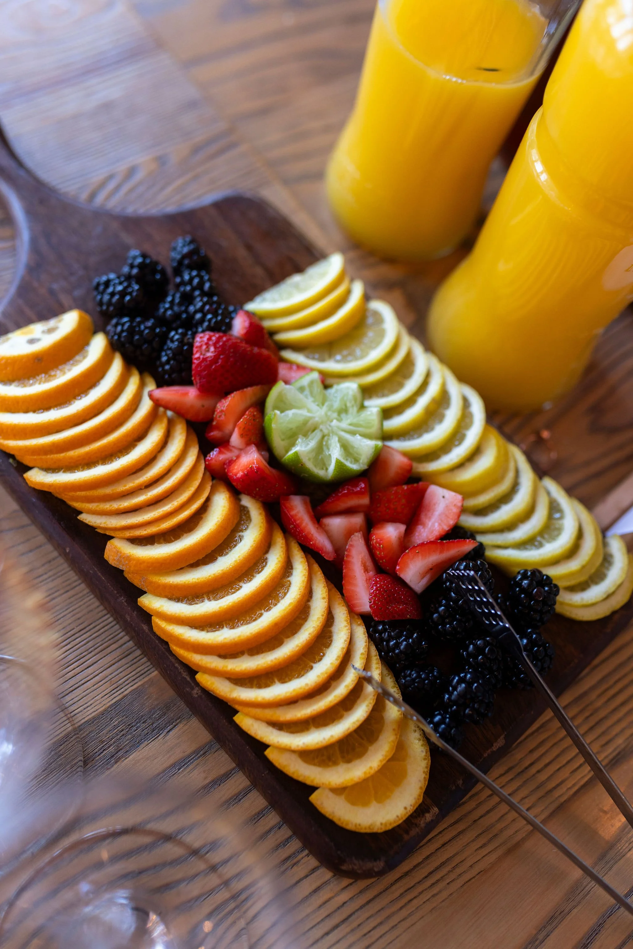A wooden serving tray with sliced oranges, strawberries, blackberries, and limes, accompanied by two glasses of orange juice on a wooden table.