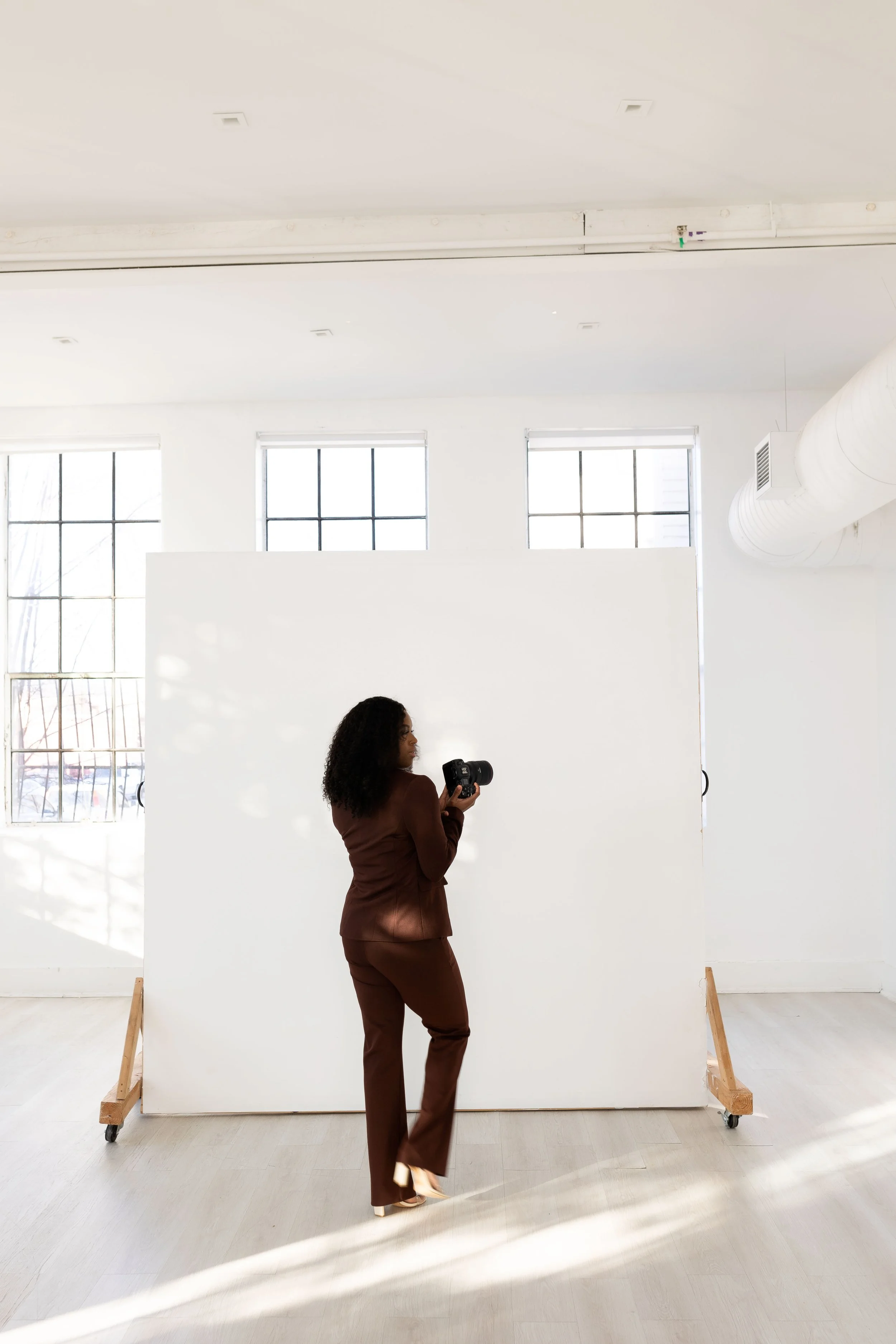 A woman with curly hair wearing a brown suit looking at a camera in a bright, minimalistic room with large windows and a white backdrop.