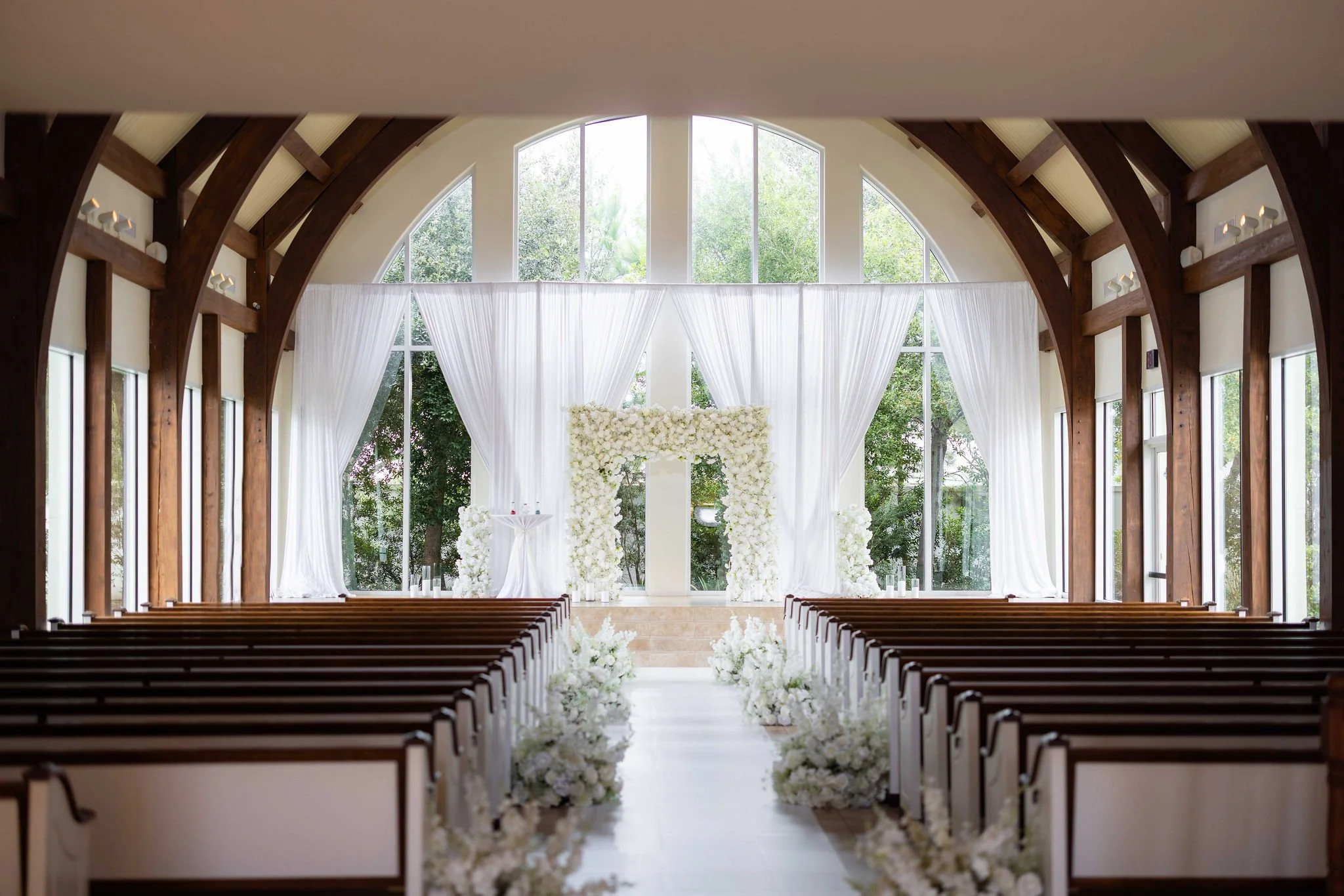 Interior of a church decorated for a wedding, with rows of wooden pews, white drapes, floral arrangements, and large windows showing greenery outside.
