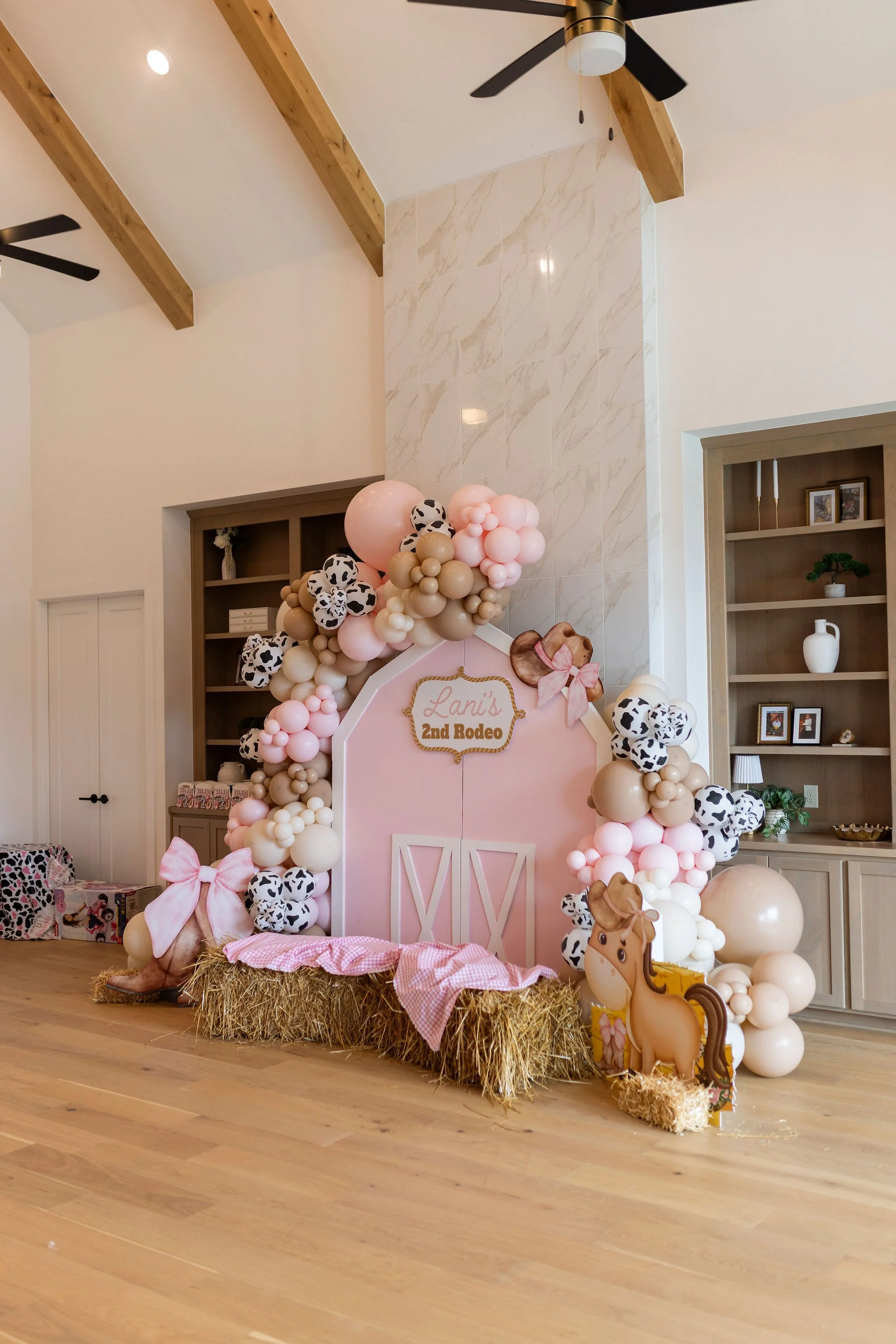 Decorative setup for a child's birthday with a pink backdrop, balloon arch with cow print and pink balloons, hay bale base, pink bows, a wooden horse figure, and a sign reading 'Lani's 2nd Rodeo' in a room with wooden floors and shelves.