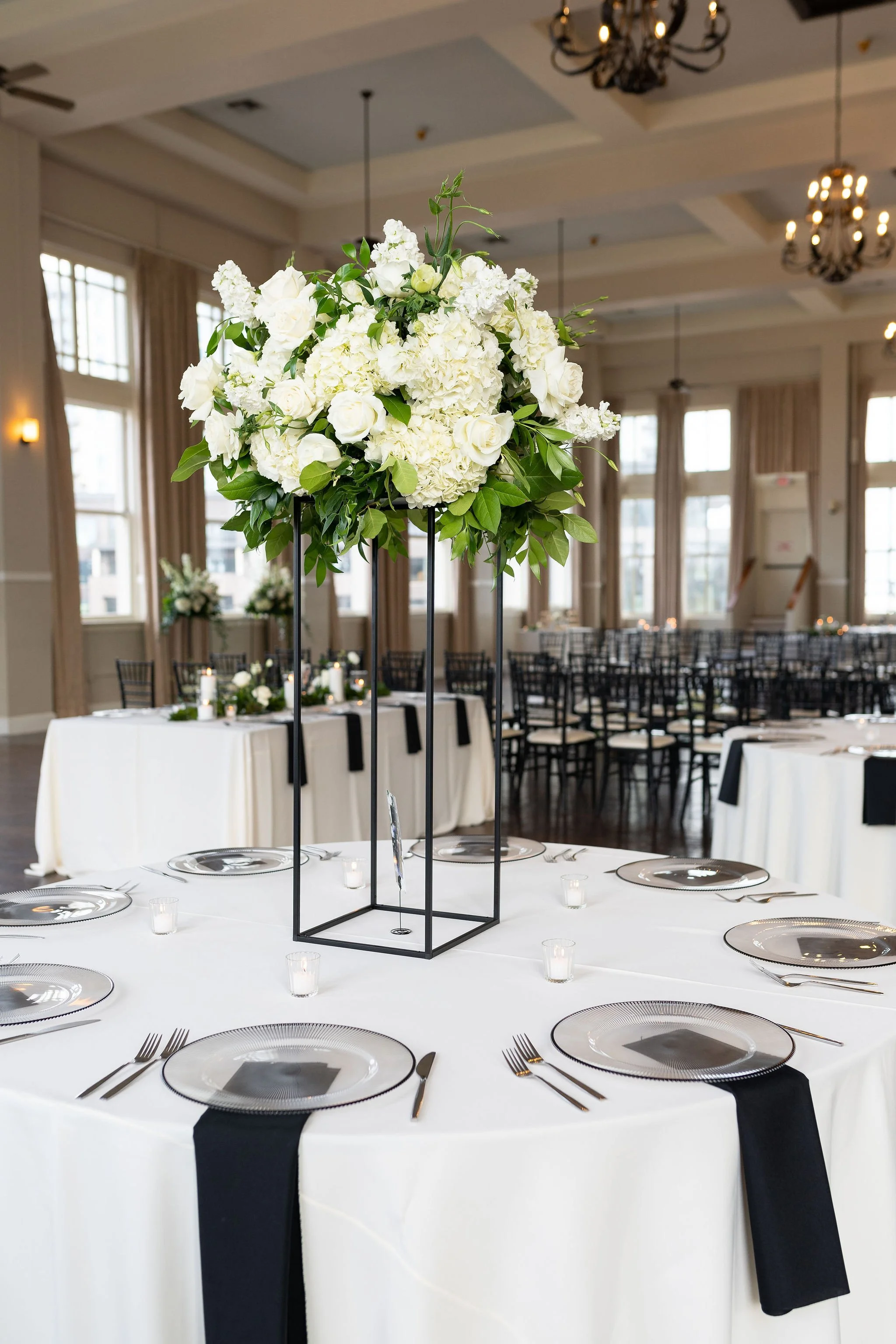 A formal dining setup with a white tablecloth, black napkins, and transparent plates, featuring a tall floral centerpiece of white roses, hydrangeas, and greenery. The room has large windows, beige curtains, candelabra chandeliers, and multiple table