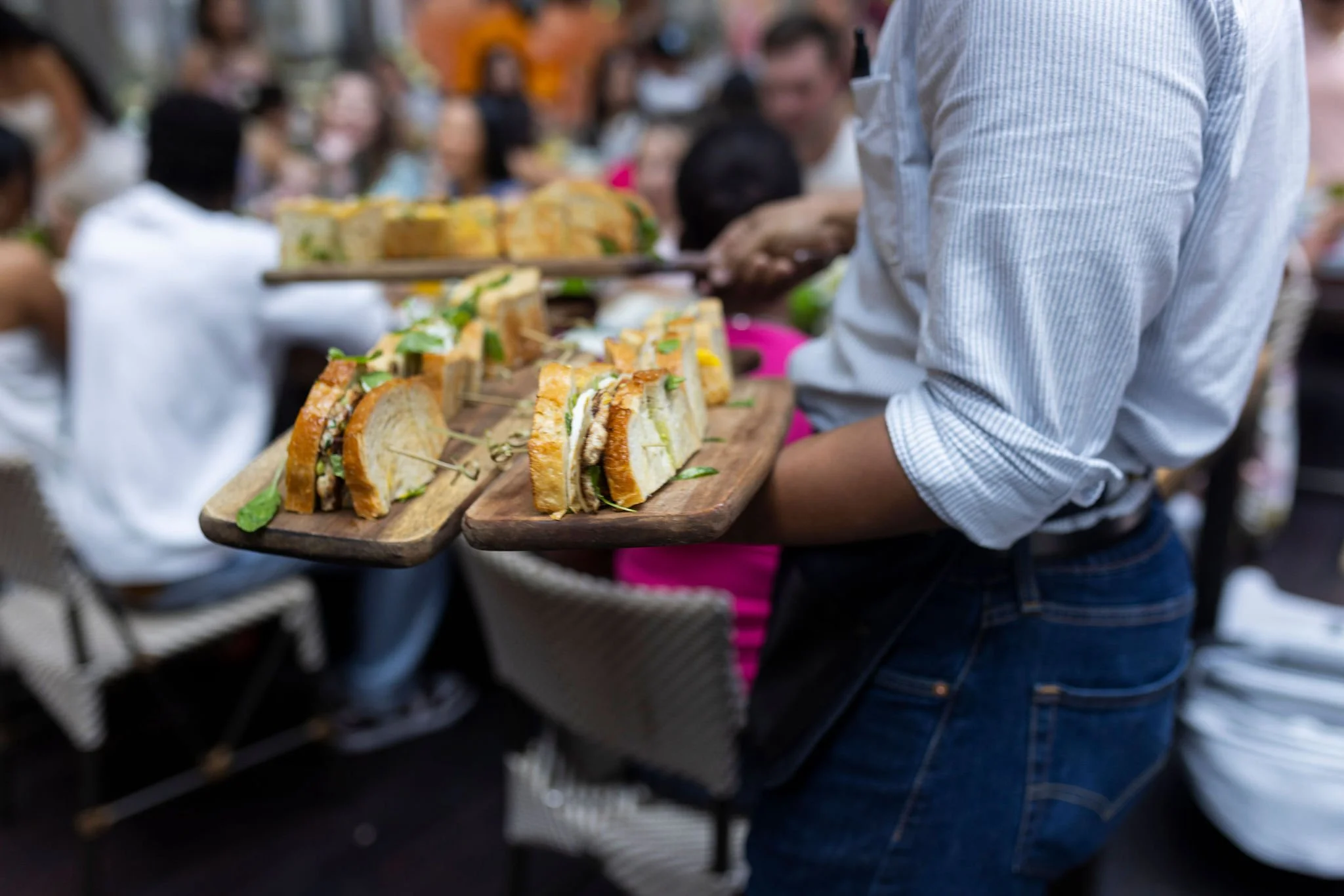 A person holding a wooden tray filled with sandwiches at an outdoor gathering or event.