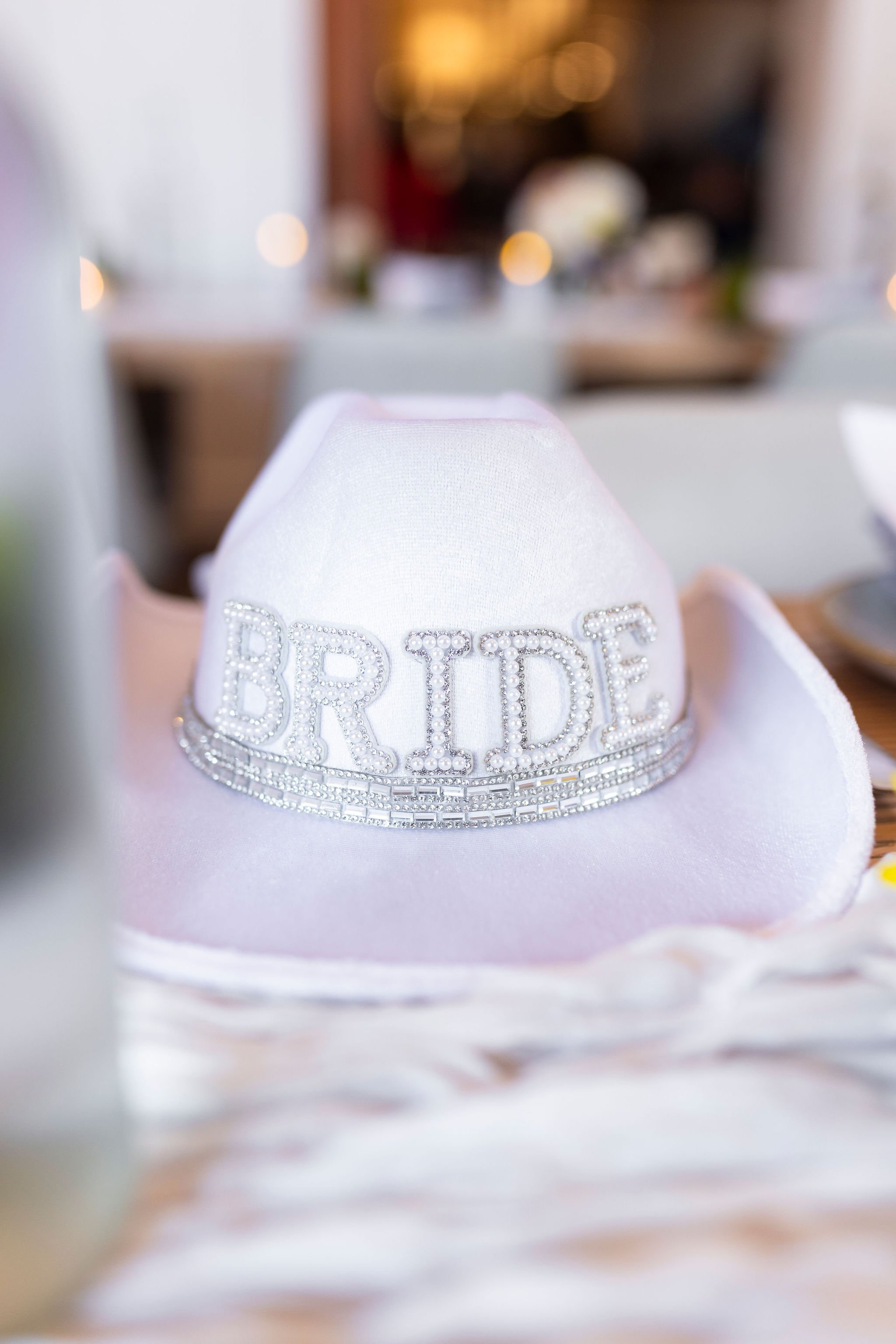 A close-up view of a white hat with the word 'BRIDE' written in rhinestones, placed on a soft fabric surface with a blurred background.