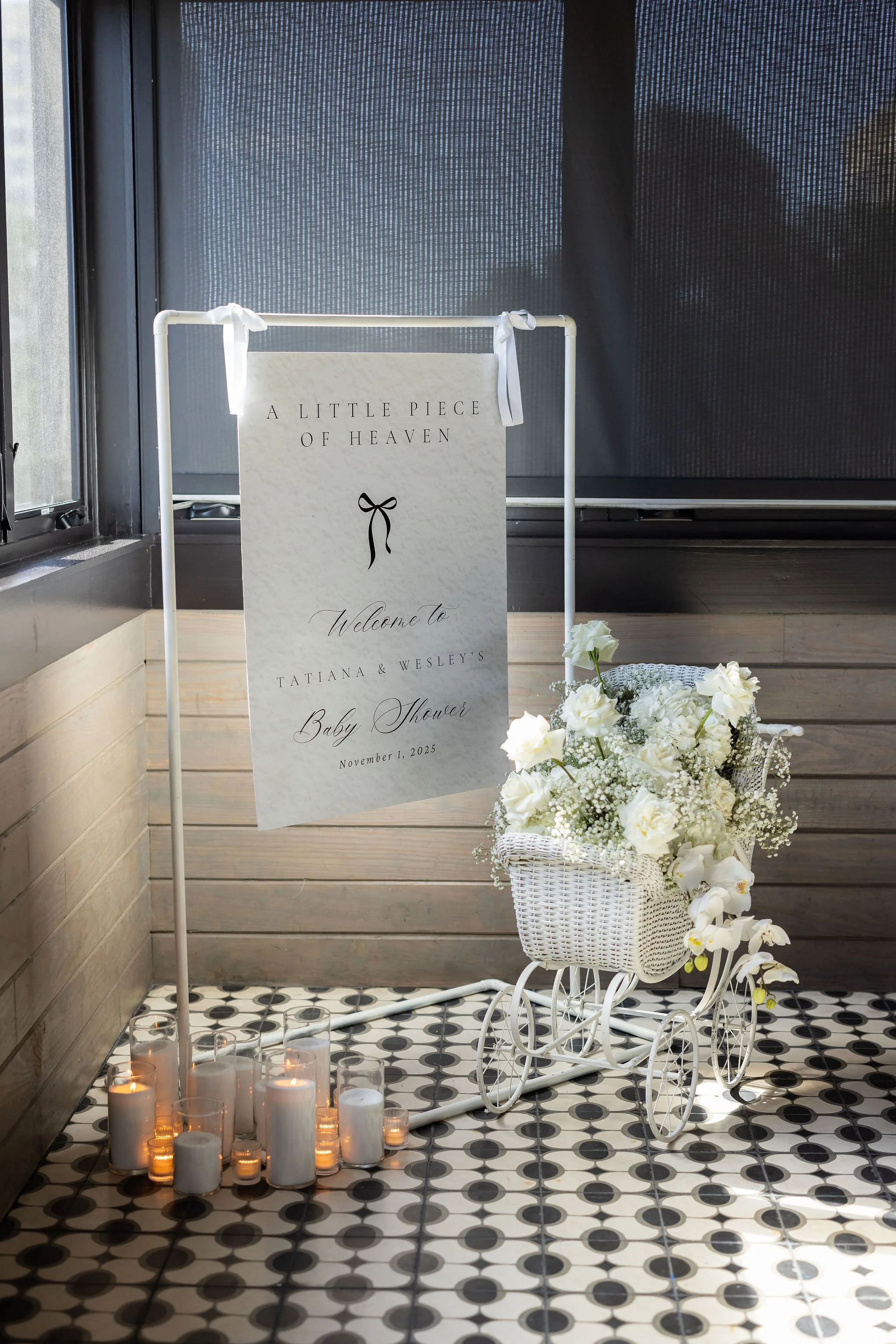 A decorative baby shower display with a white tricycle planter filled with white roses and orchids, a sign that reads 'A little piece of heaven, welcome to Tatiana & Wesley's baby shower, November 1, 2025,' and candles in glass jars on a patterned black and white tile floor.