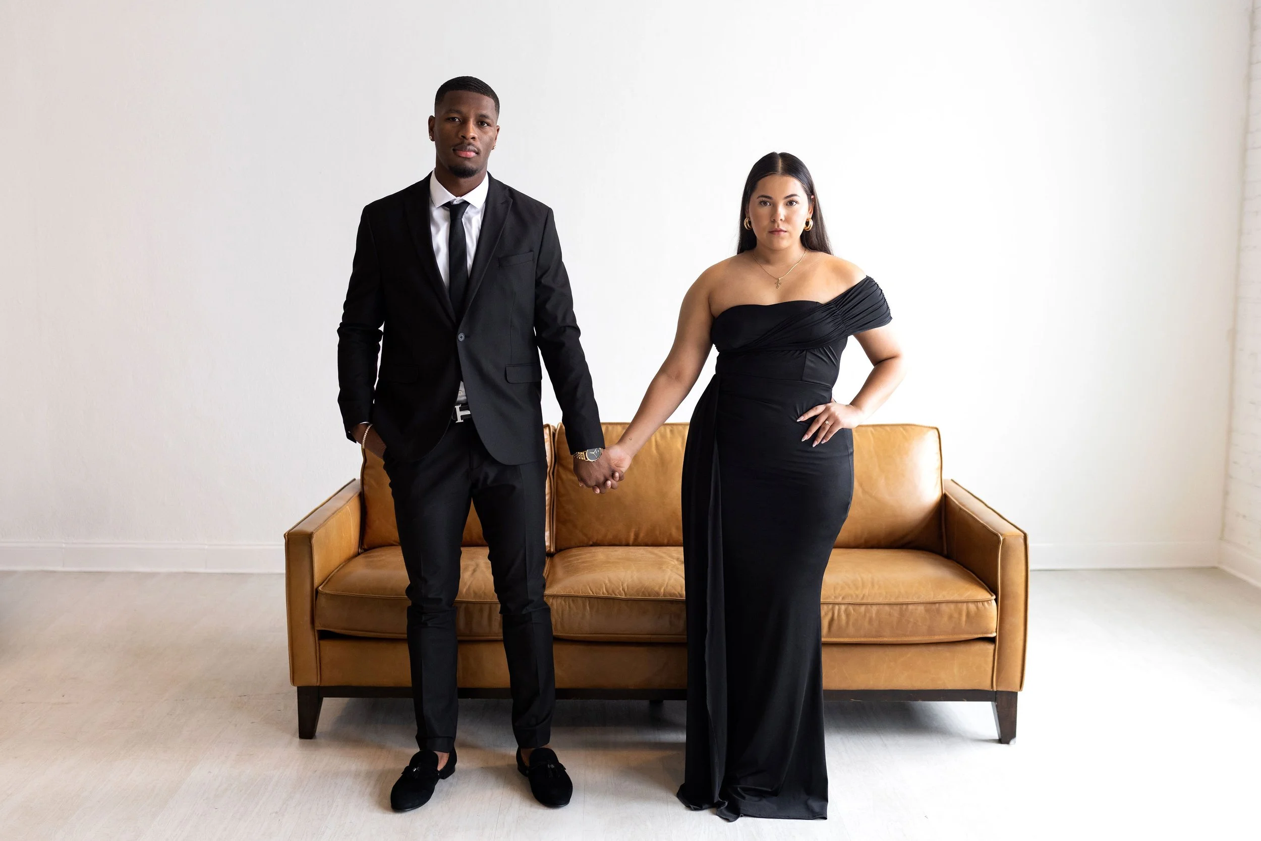 A man and woman holding hands, standing in front of a mid-century modern sofa in a minimalist room with white walls.