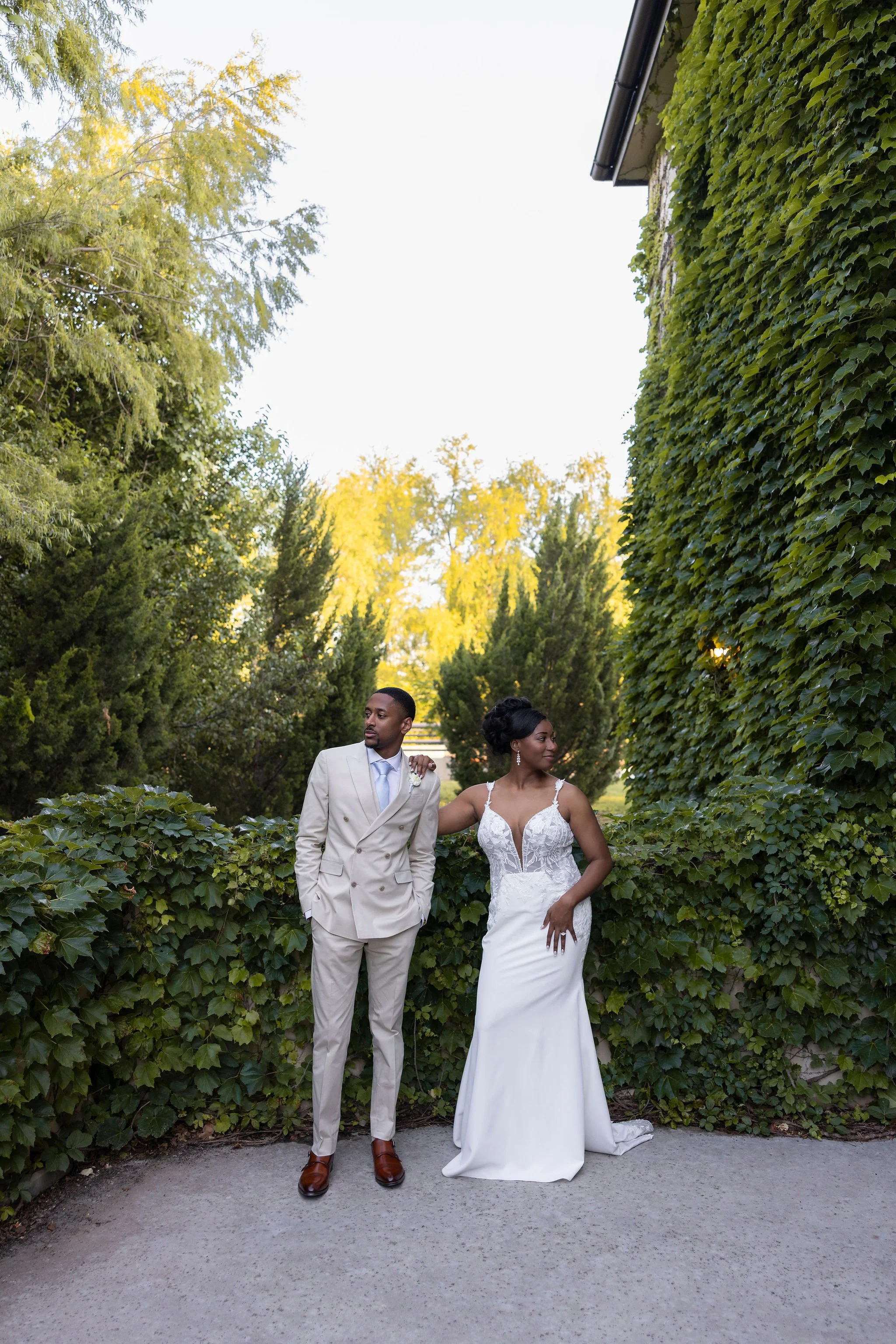 A bride and groom standing outdoors in a garden surrounded by lush green ivy-covered walls and trees, with the groom in a light-colored suit and the bride in a white wedding gown.