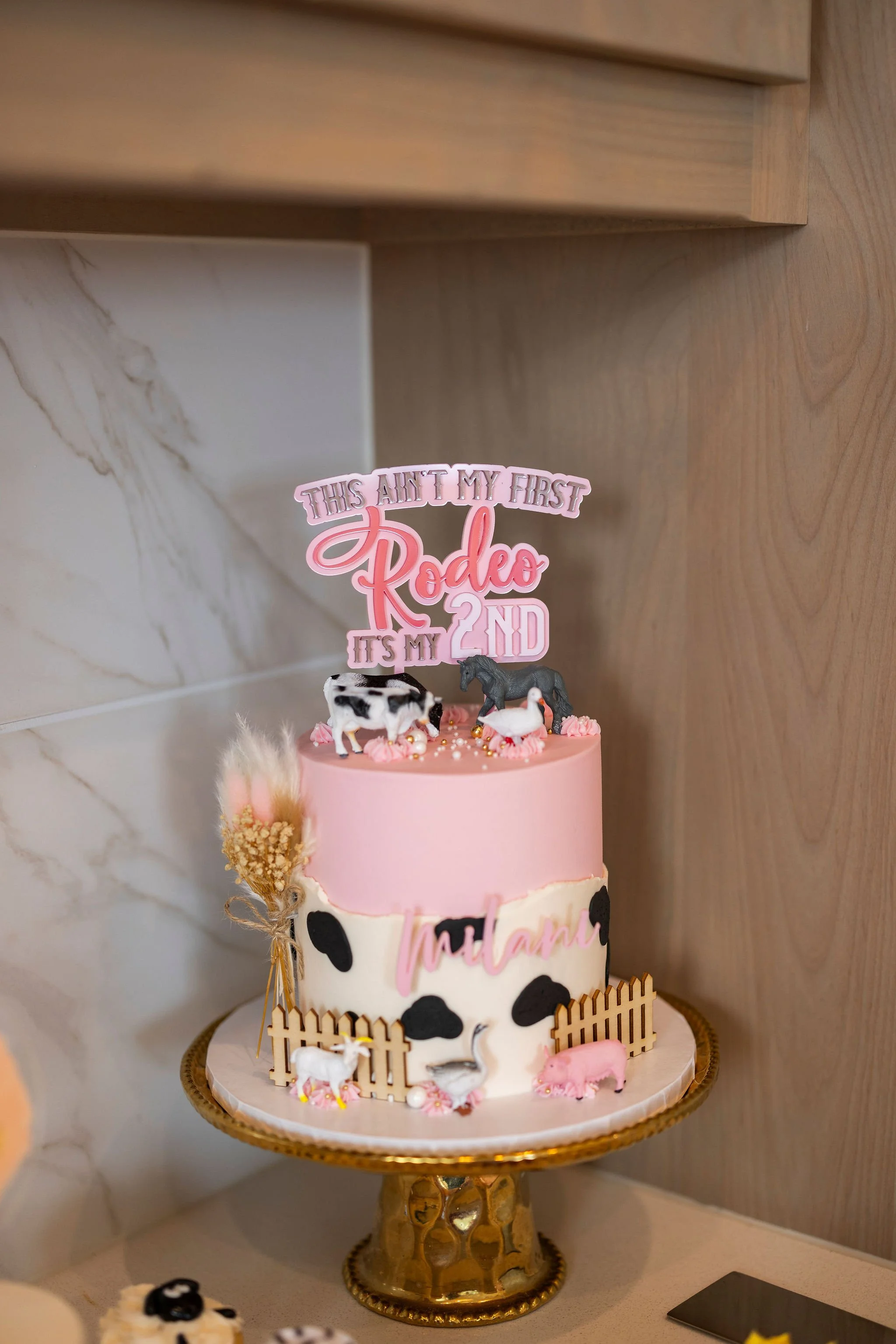 A two-tiered cake decorated with cow print patterns, pink frosting, small animal figurines, a fenced border, and a pink cake topper that reads "This ain't my first rodeo, it's my 2nd."