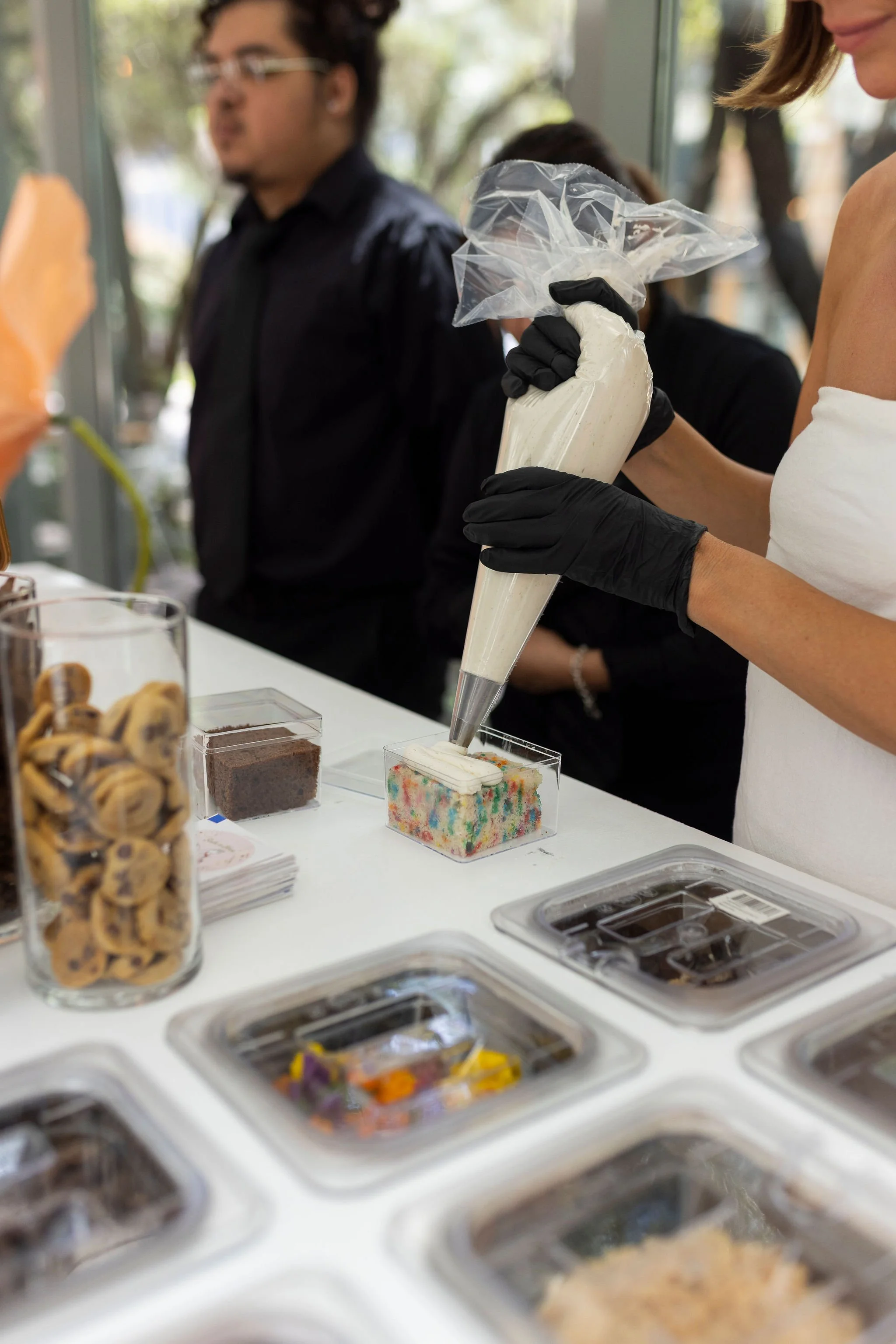 A person wearing a white tank top and black gloves filling a clear plastic bag with rainbow-colored ice cream from a pastry bag at an ice cream shop. Several containers of toppings and desserts are on the white counter in the foreground, and another 