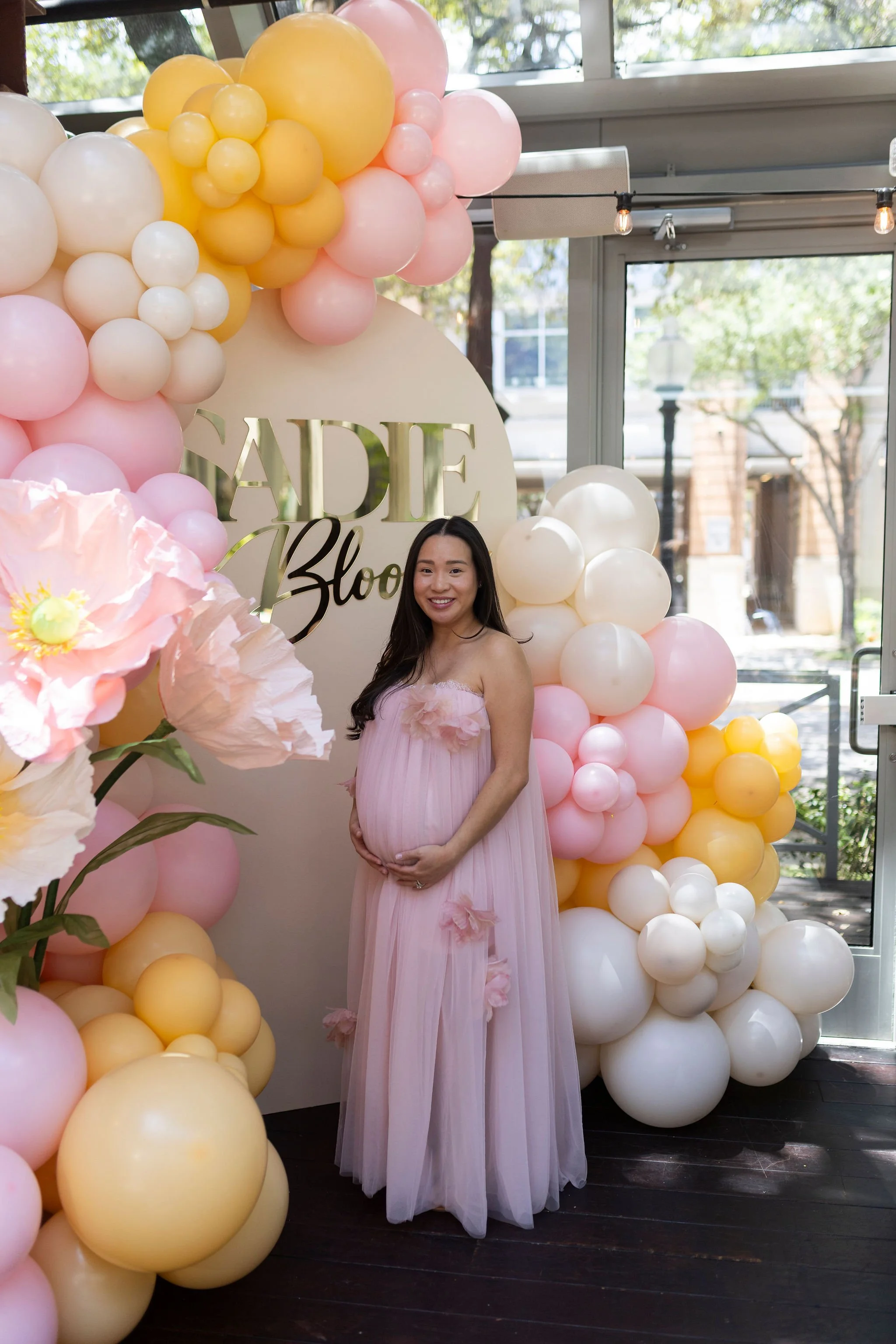 A pregnant woman in a pink dress standing in front of a pastel-colored balloon arch with a sign that reads 'Baby'. Sunlight coming through windows in the background.