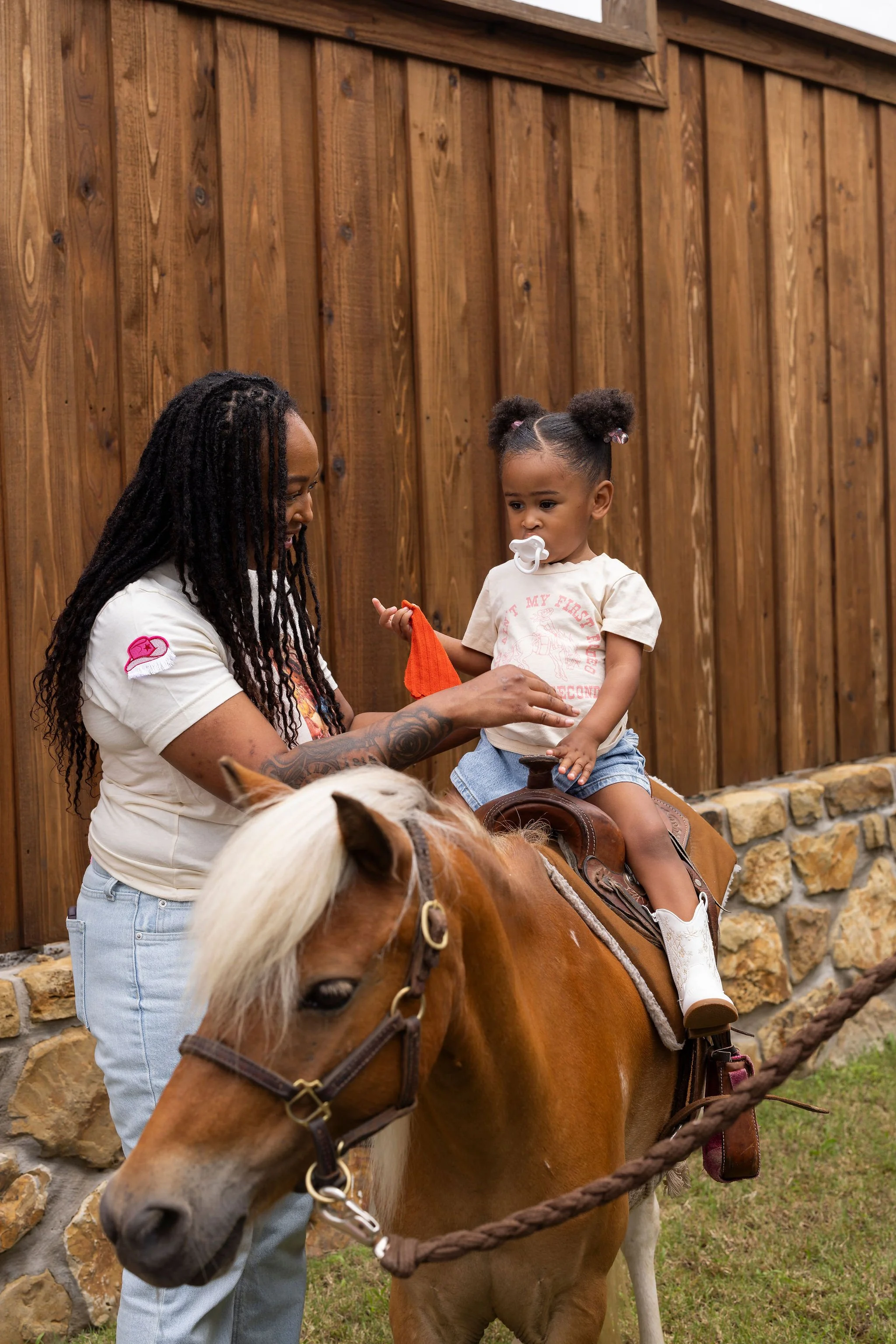 A woman with dreadlocks and tattoos on her arms is helping a young girl sit on a brown and white pony. The girl has her hair styled in two puffs and is holding a small carrot, with a pacifier in her mouth. They are outside near a wooden fence with a 
