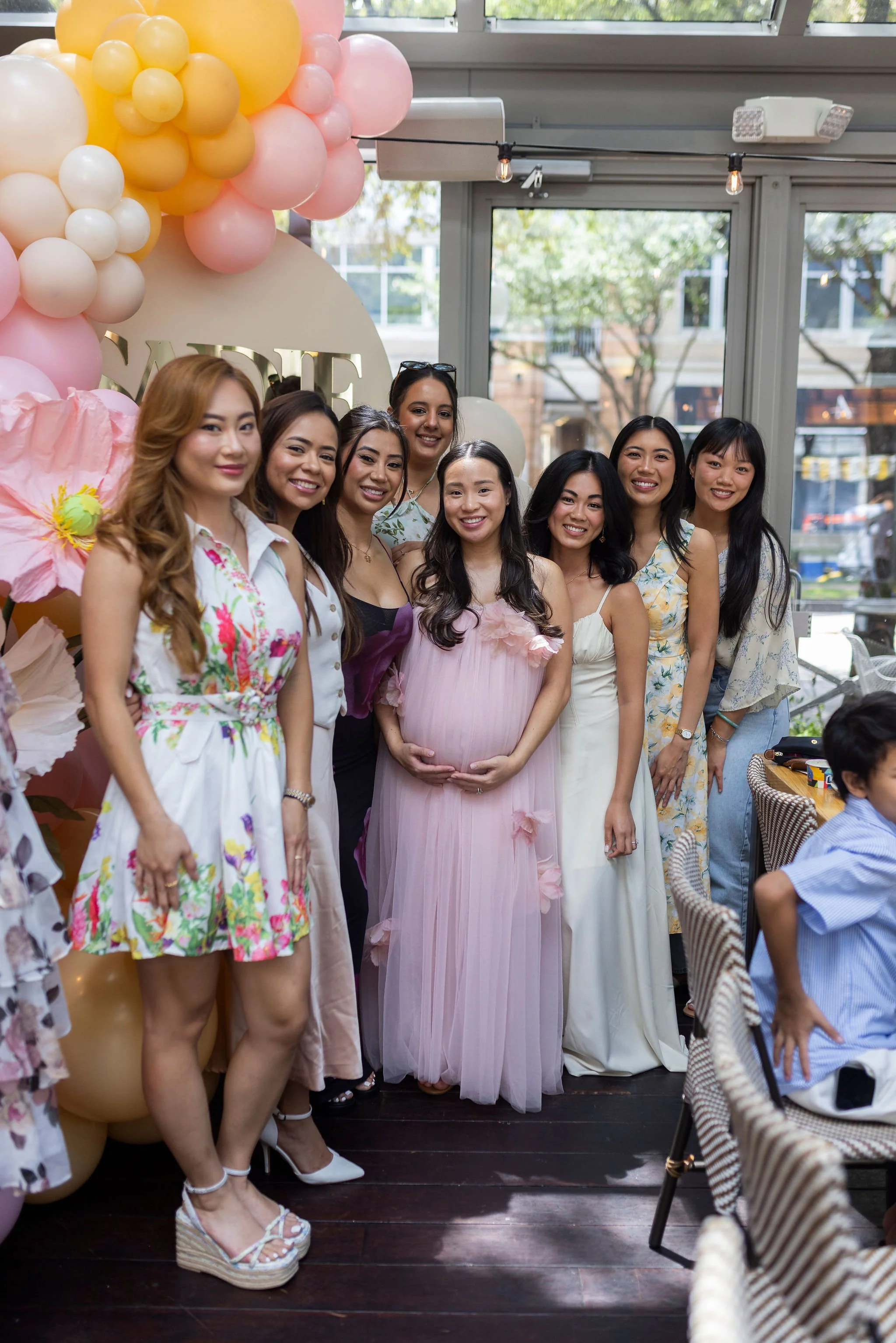Women gathered at a celebration, with a woman in the center wearing a pink dress holding her pregnant belly. The background includes pink and yellow balloons, a window, and outdoor scenery.