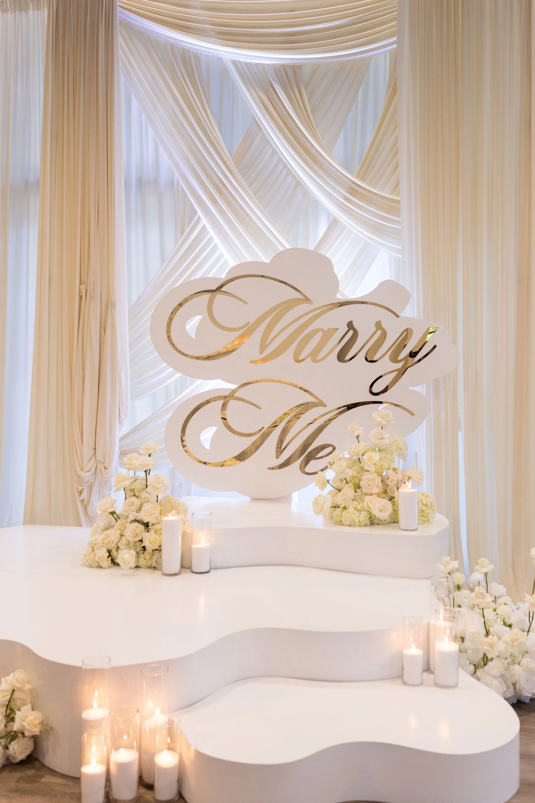 Elegant wedding or event cake table decorated with white flowers, tall white candles in glass holders, and a backdrop of flowing white fabric with soft lighting. A decorative sign or banner with gold accents reads "Marry Me".
