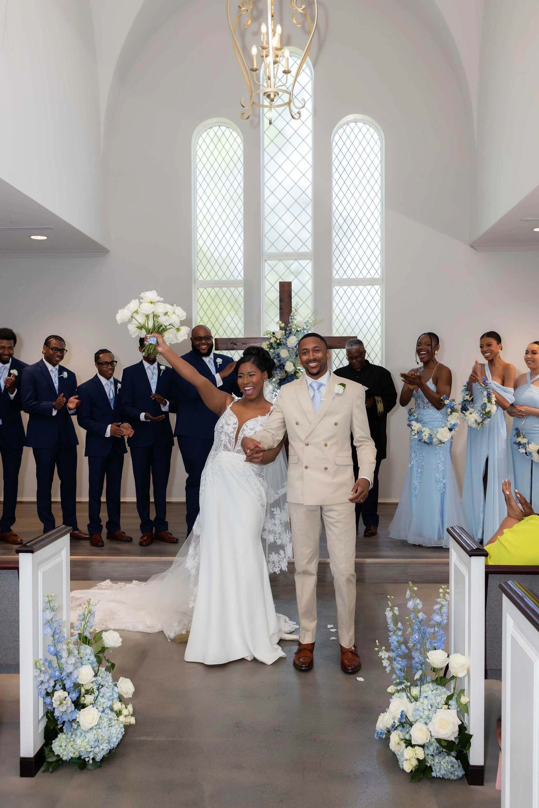 A newlywed couple holding hands and smiling at the altar during a wedding ceremony inside a chapel with tall windows, a hanging chandelier, and floral decorations.