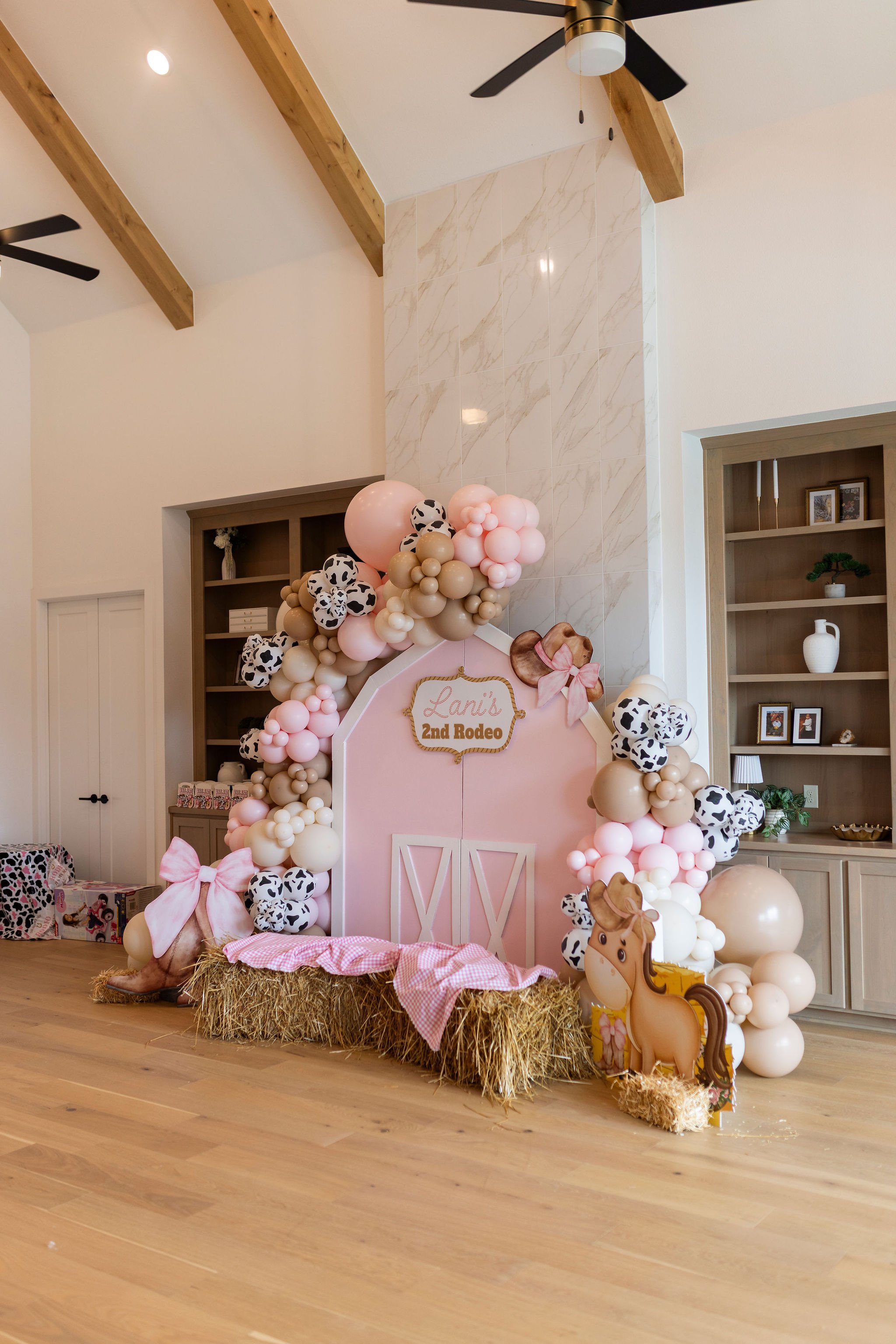 Decorative backdrop with balloons and a sign for a child's birthday, set up for a farm or cowboy theme with a wooden horse figure, hay bales, and pink bows, in a room with wooden floors and ceiling with beams.