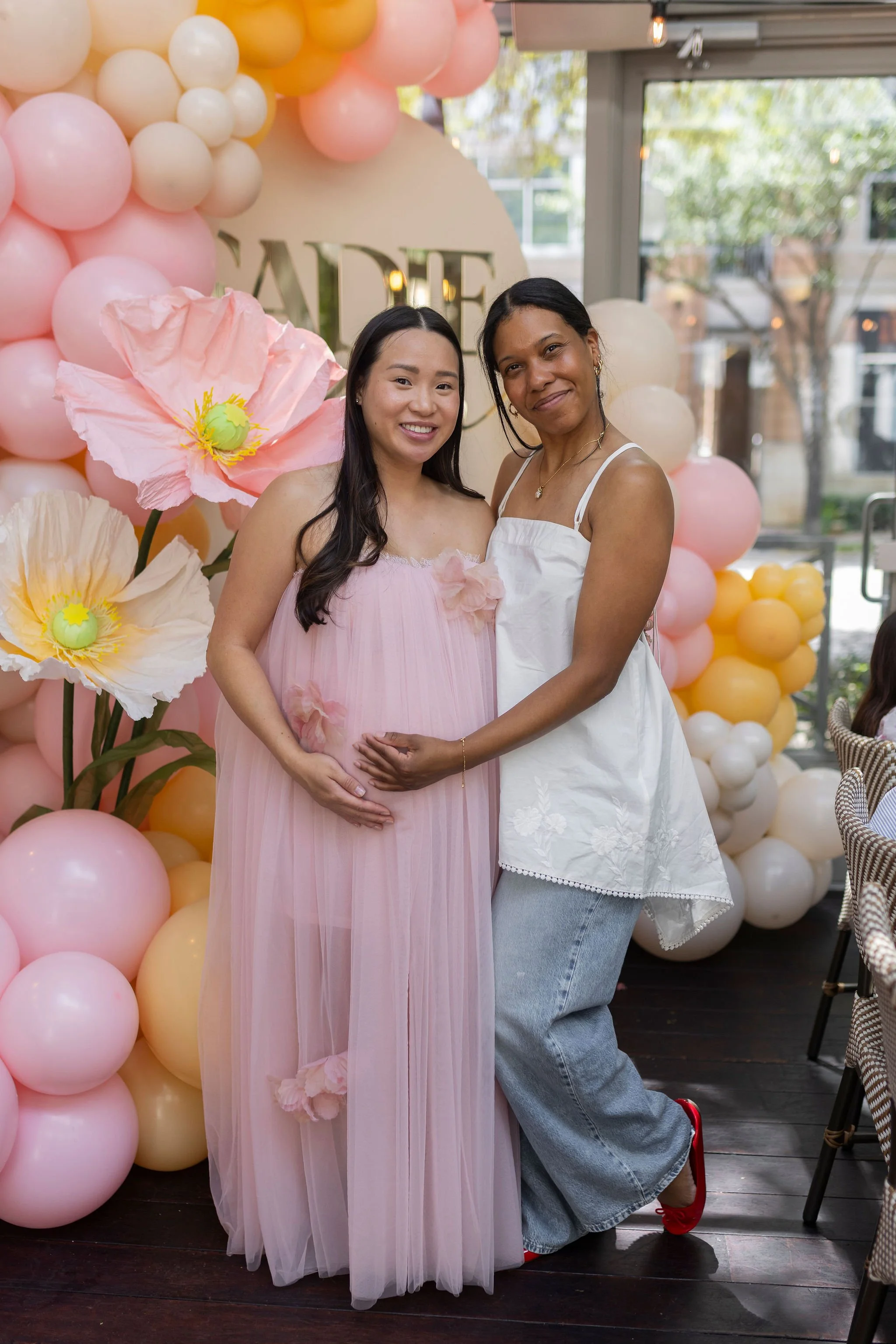 Two women standing in front of a pink and yellow balloon decoration at a baby shower, with a large paper flower and a sign that spells 'BABY' in the background.