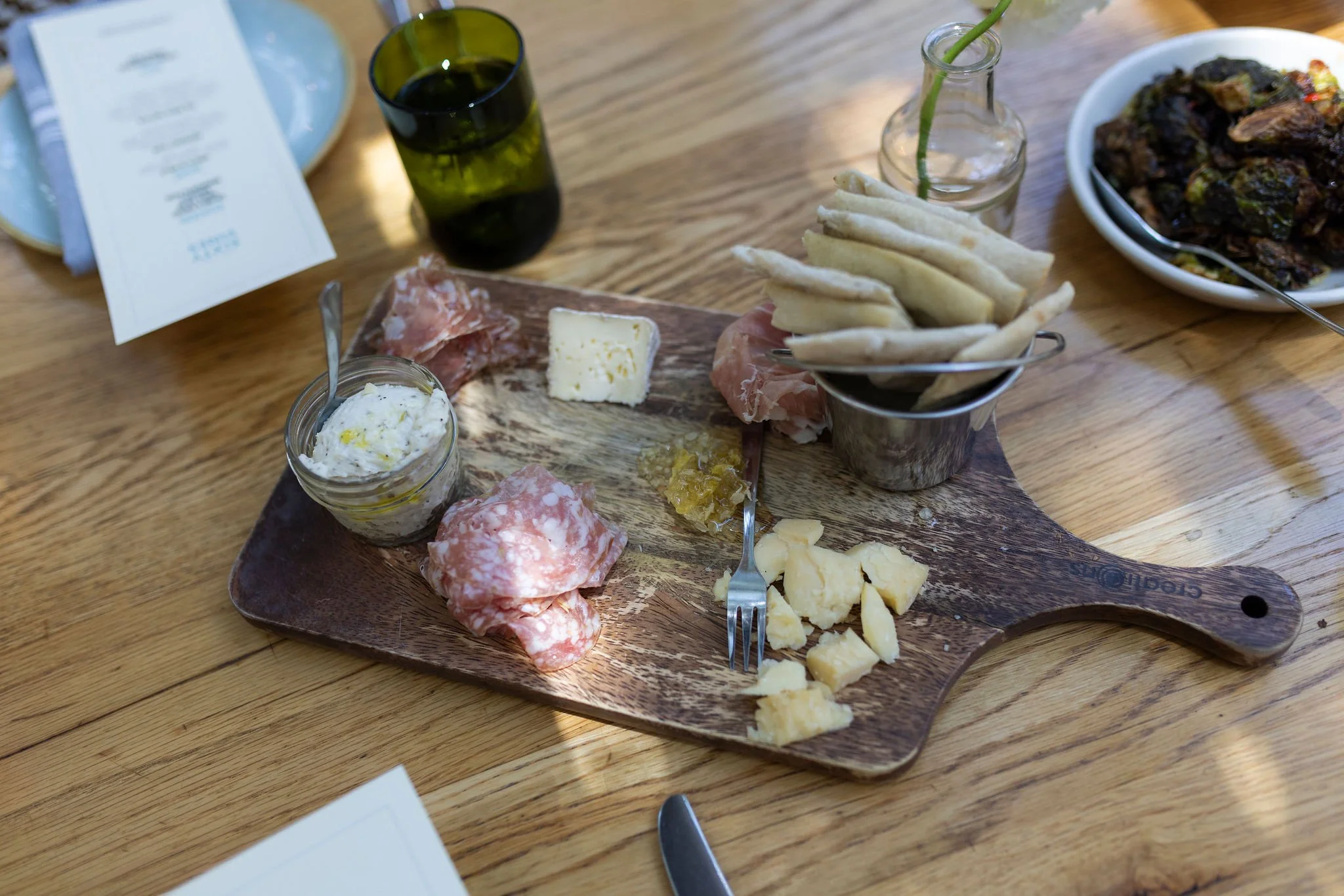 A wooden platter of assorted cheeses, meats, and condiments, with a small jar of creamy spread, slices of cured meat, cheeses, pita bread, and a knife, on a wooden table with a menu and a glass of dark beverage in the background.