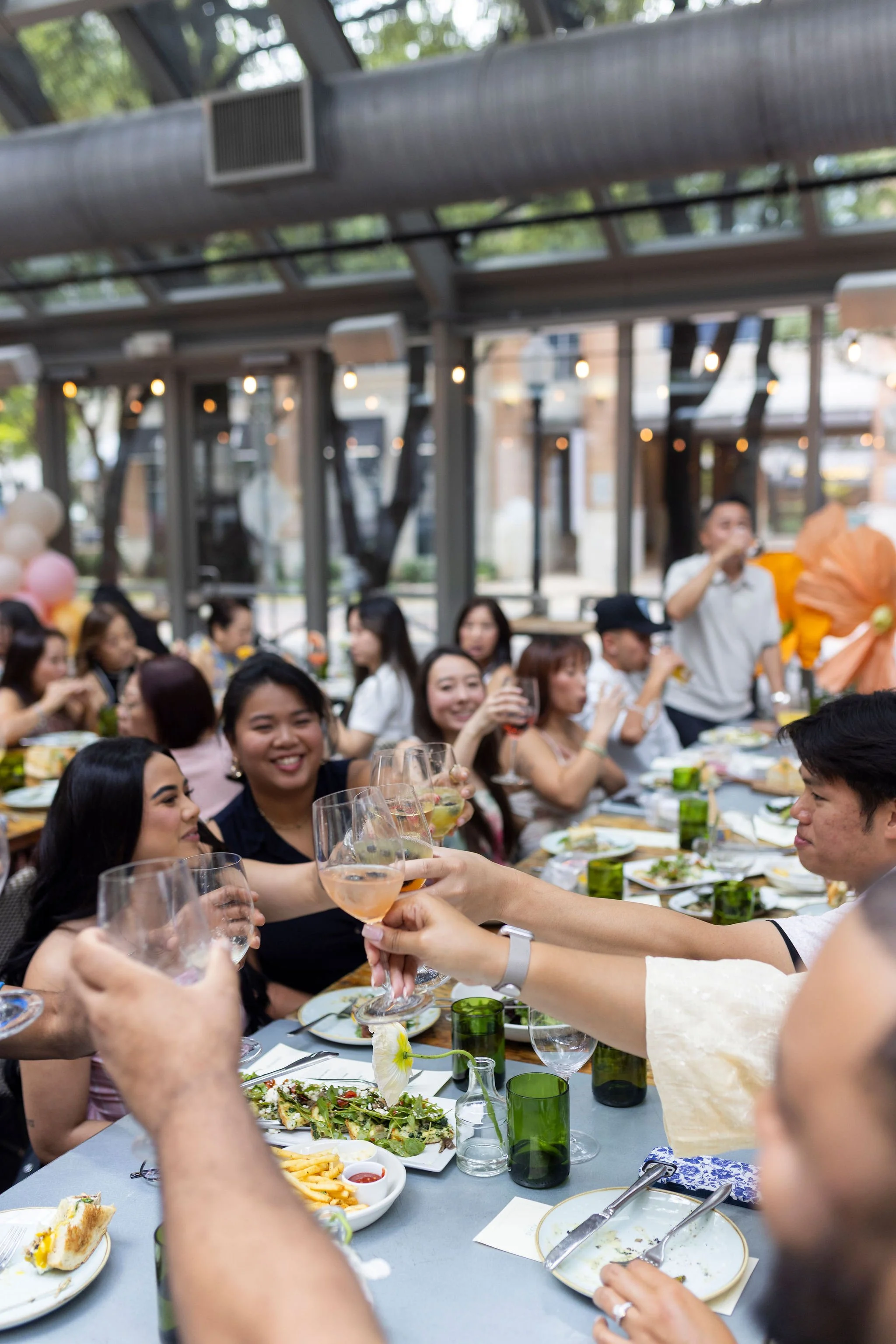 People celebrating at a social gathering with food and drinks at a long table in a covered outdoor setting.
