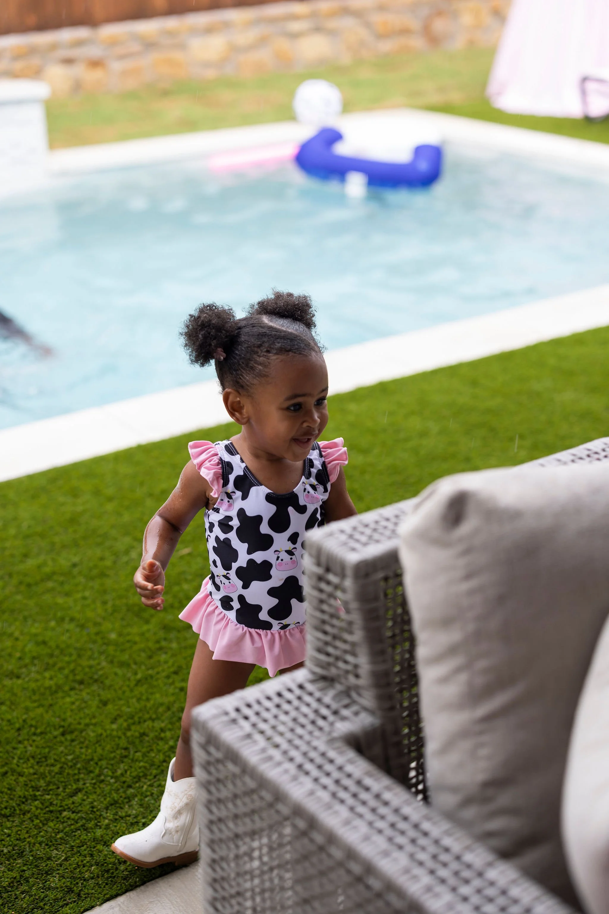 A young girl with curly hair styled in puffy pigtails, wearing a cow-print top with pink ruffle sleeves, a pink skirt, and white cowboy boots, is playing outdoors near a swimming pool with a stone wall in the background and a wicker basket in the for