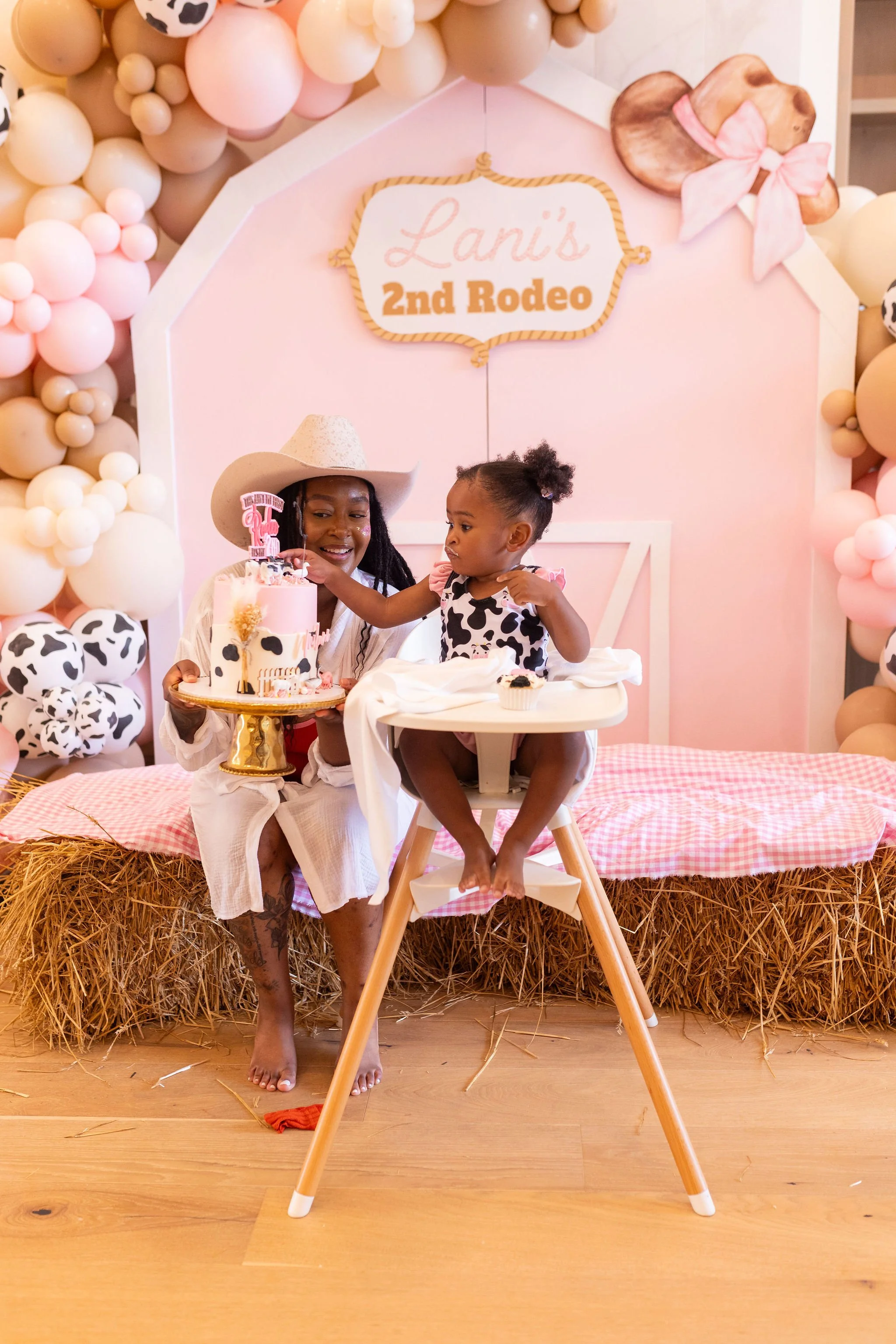 A woman in white shorts and a cowboy hat sits barefoot on a surface covered with hay, holding a pink cow-themed birthday cake with candles. Next to her, a young girl in a cow-print dress sits in a high chair, reaching for the cake. Behind them is a p