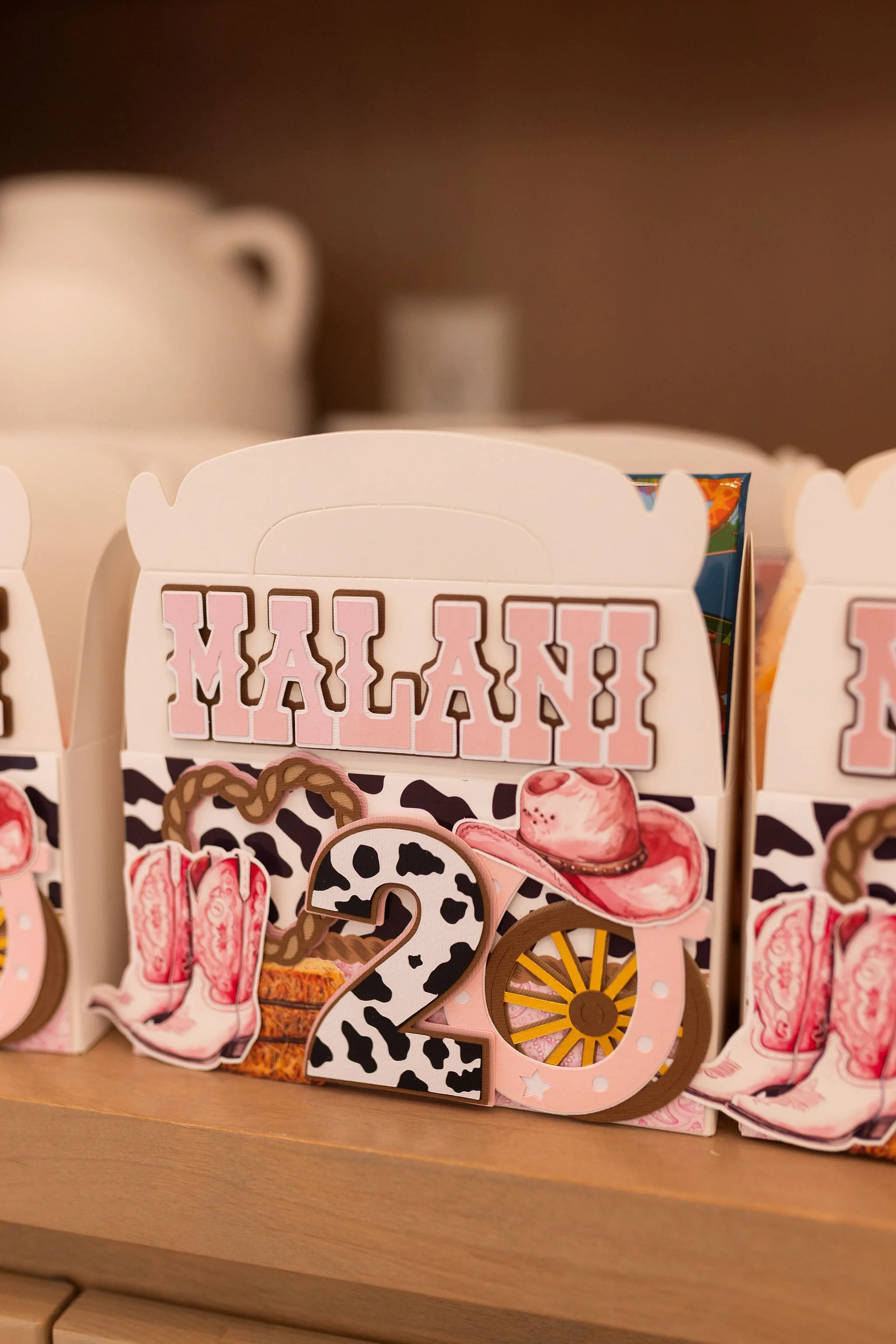 Decorative items forming the phrase 'MALANI' with various pink, black, and cow-print designs, including a cowboy hat, shoes, and wheels, on a wooden surface.