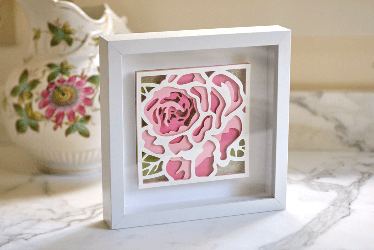 Decorative white framed 3D pink and green floral artwork on a white marble surface with a floral patterned pitcher in the background.