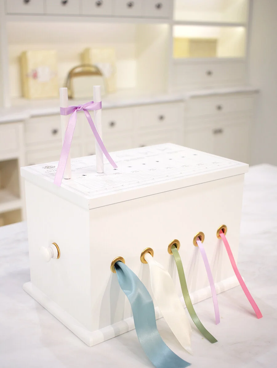 A white gift box with colorful satin ribbons and a small decorated cross on top, set in a brightly lit room with white cabinets and drawers in the background.