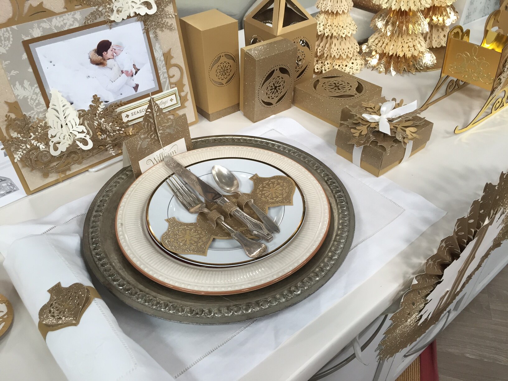 Decorative holiday table setting with gold and white accents, including a photo frame, wrapped gifts, Christmas trees, and ornamental plates with cutlery.