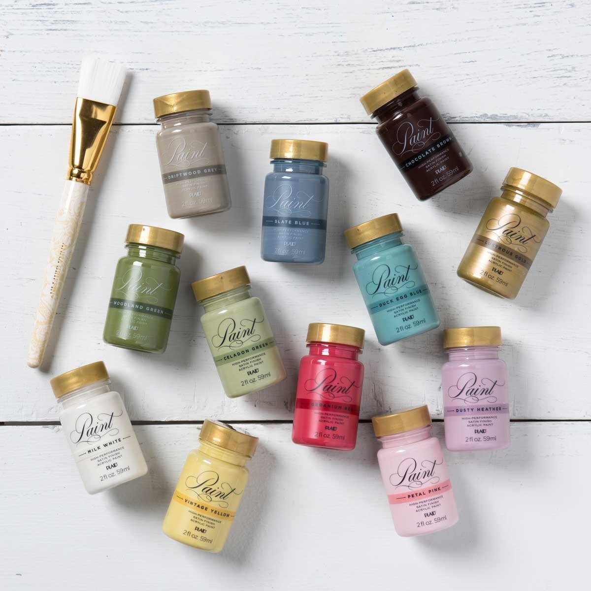 Collection of small jars of colored paint with gold lids, arranged on a white wooden surface, including colors such as white, yellow, pink, blue, green, black, brown, and purple, with a paintbrush on the side.