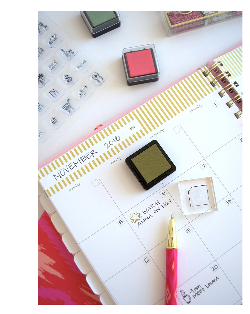 A desk calendar open to November 2018, with handwritten notes and stamps. Several ink pads and stamps are on the desk, along with a pink and gold pen. The calendar contains scheduled events including watching TV and meeting someone named Laura.