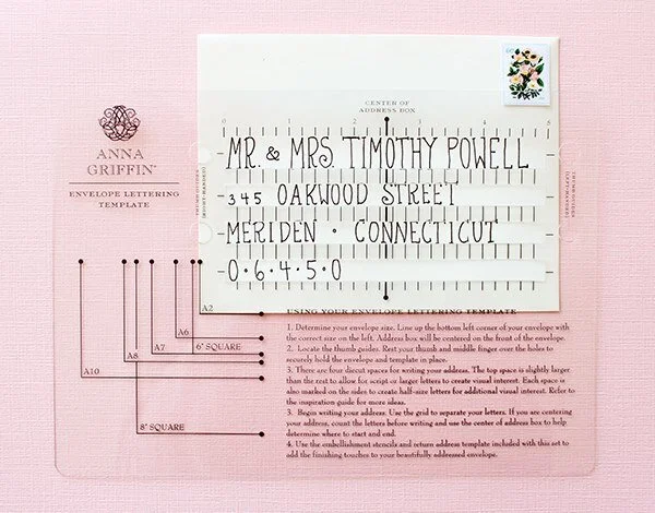 Pink envelope lettering template with handwritten address and instructions, Anna Griffin logo, and decorative floral stamp in the top right corner.