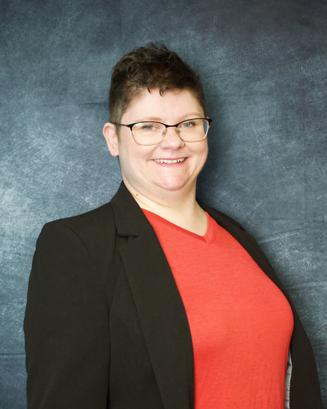 A woman with short dark hair, glasses, and a friendly smile, wearing a black blazer over a red top, standing against a gray textured background.
