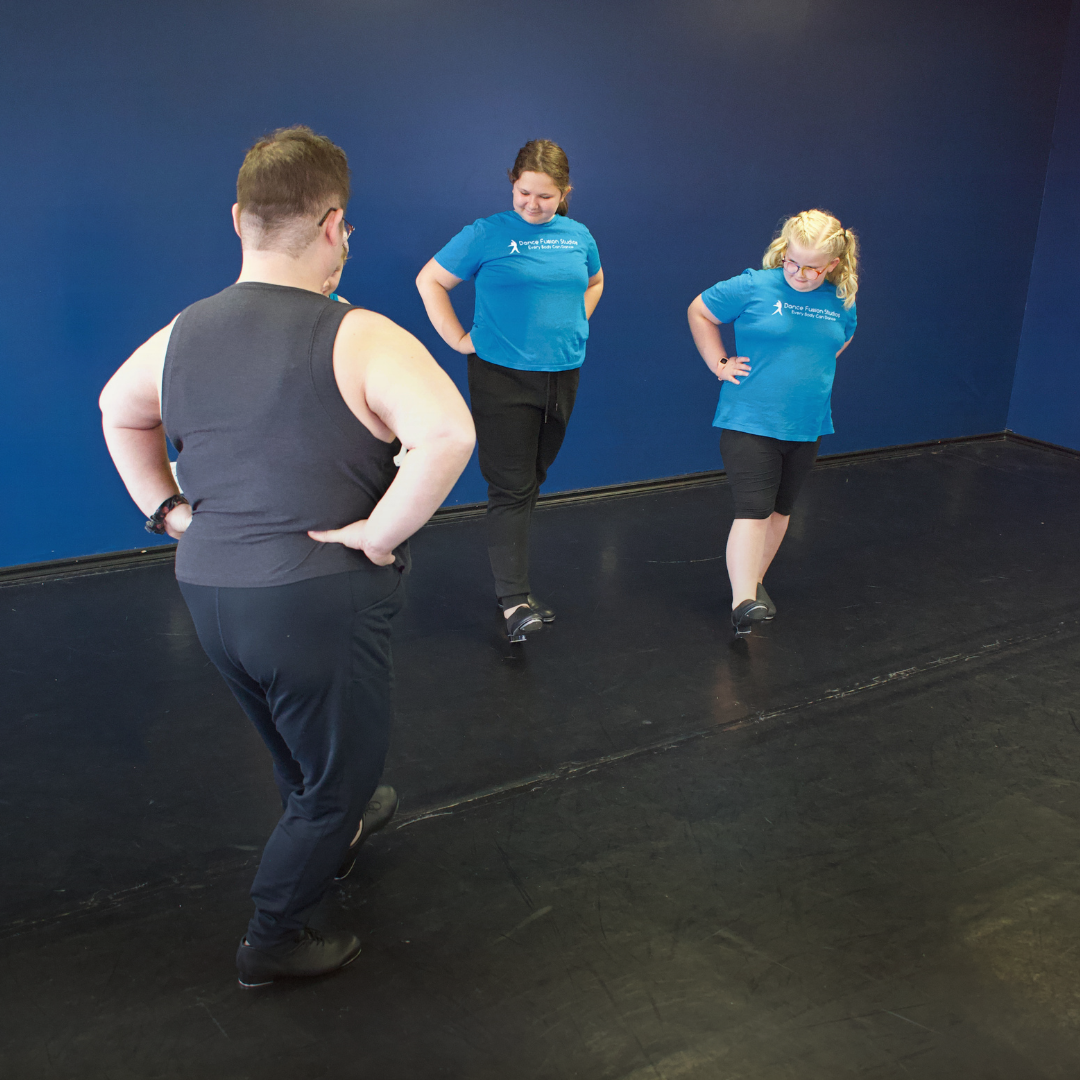 Two girls in blue shirts and one boy in a black tank top and black pants practicing a dance routine in a dance studio with a dark blue wall.