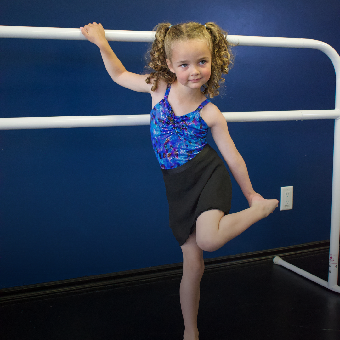 Young girl in blue dance costume practicing on a ballet barre in a dance studio with a blue wall.