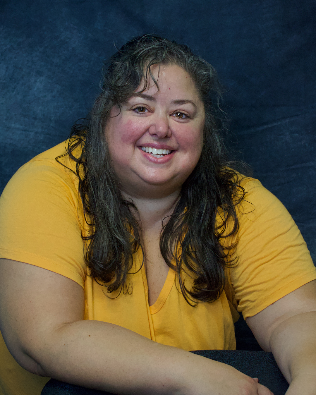 A woman with long curly hair wearing a yellow shirt, smiling and seated against a dark background.