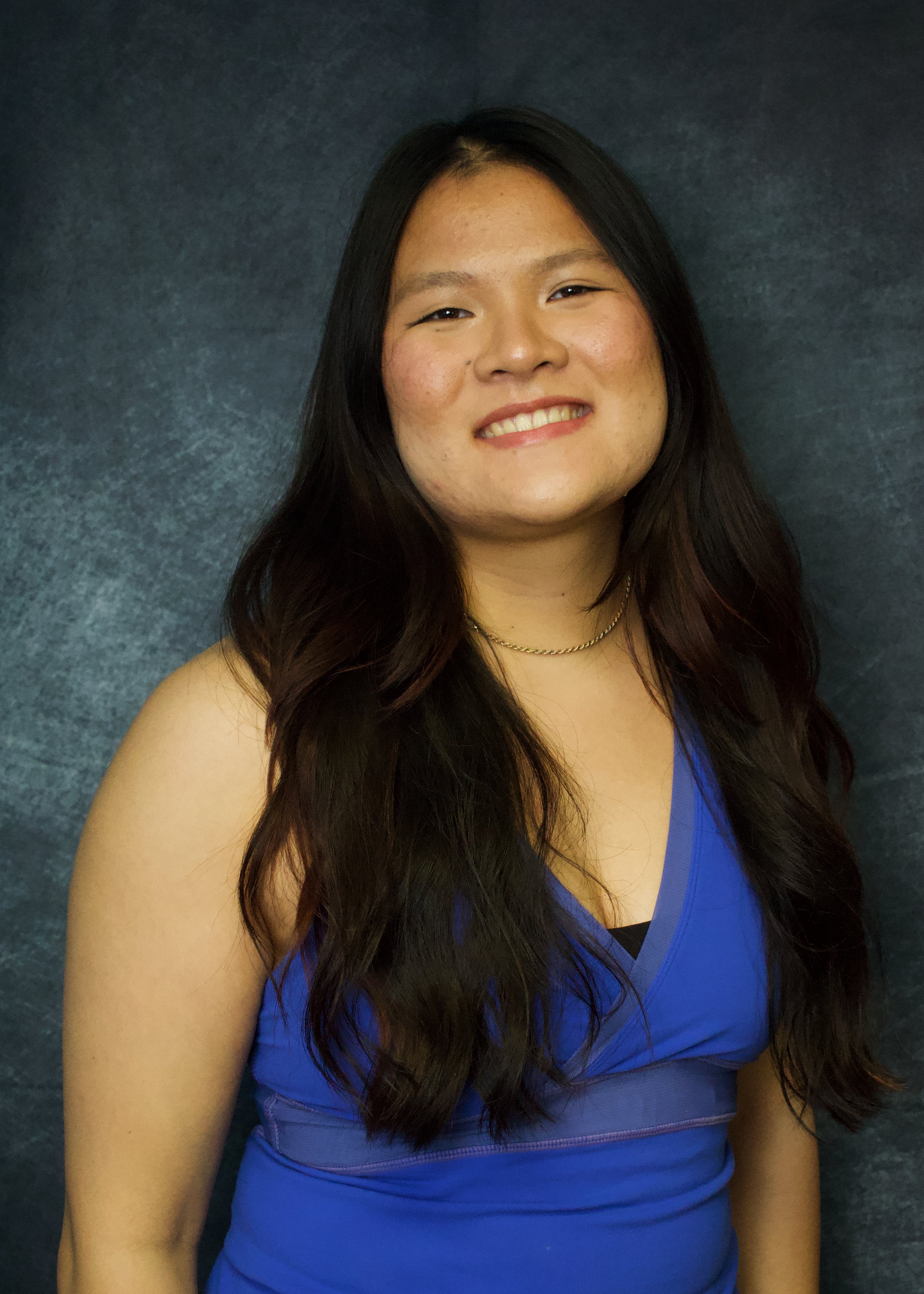 Portrait of a young woman with long dark hair, smiling, wearing a blue sleeveless top and a gold necklace, against a dark textured background.