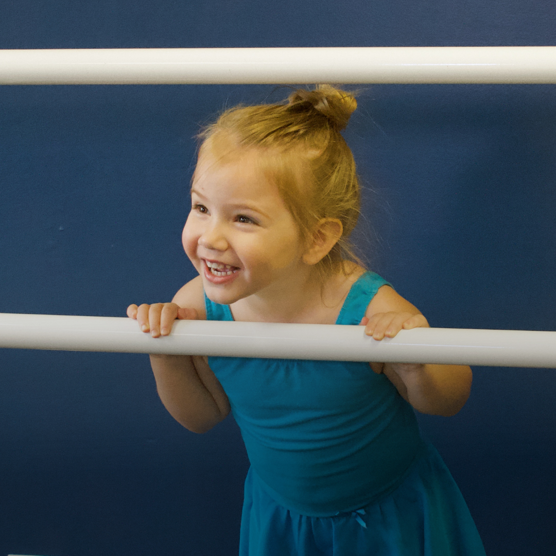 A young girl with blonde hair tied in a bun, wearing a blue outfit, smiling and holding onto a white horizontal bar against a dark blue wall.