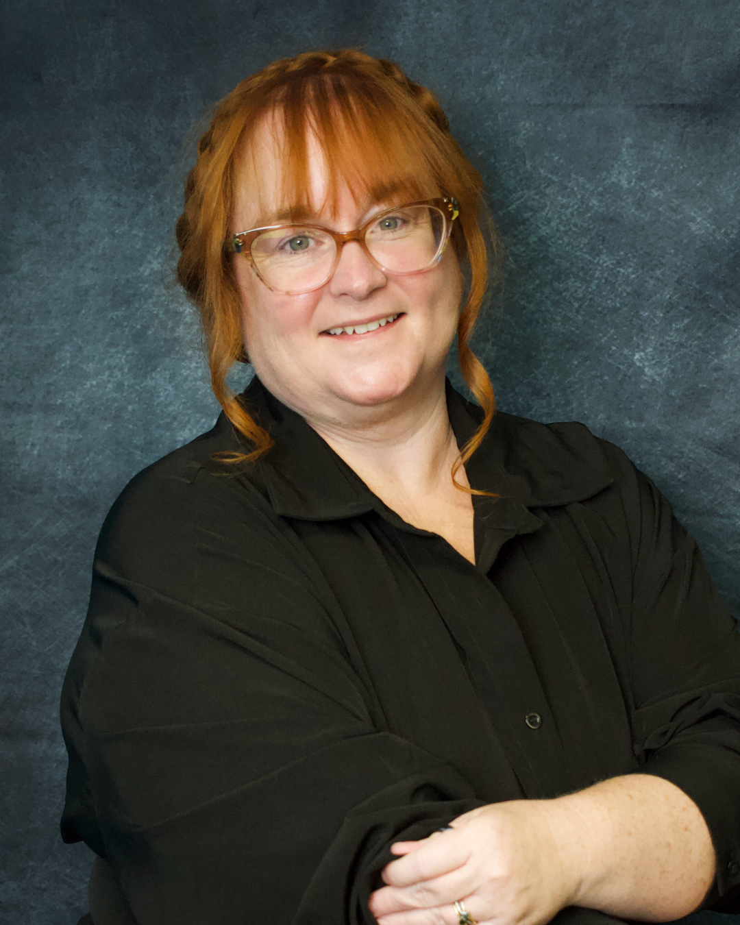 A woman with red hair, glasses, wearing a black shirt, smiling with crossed arms against a textured dark blue background.
