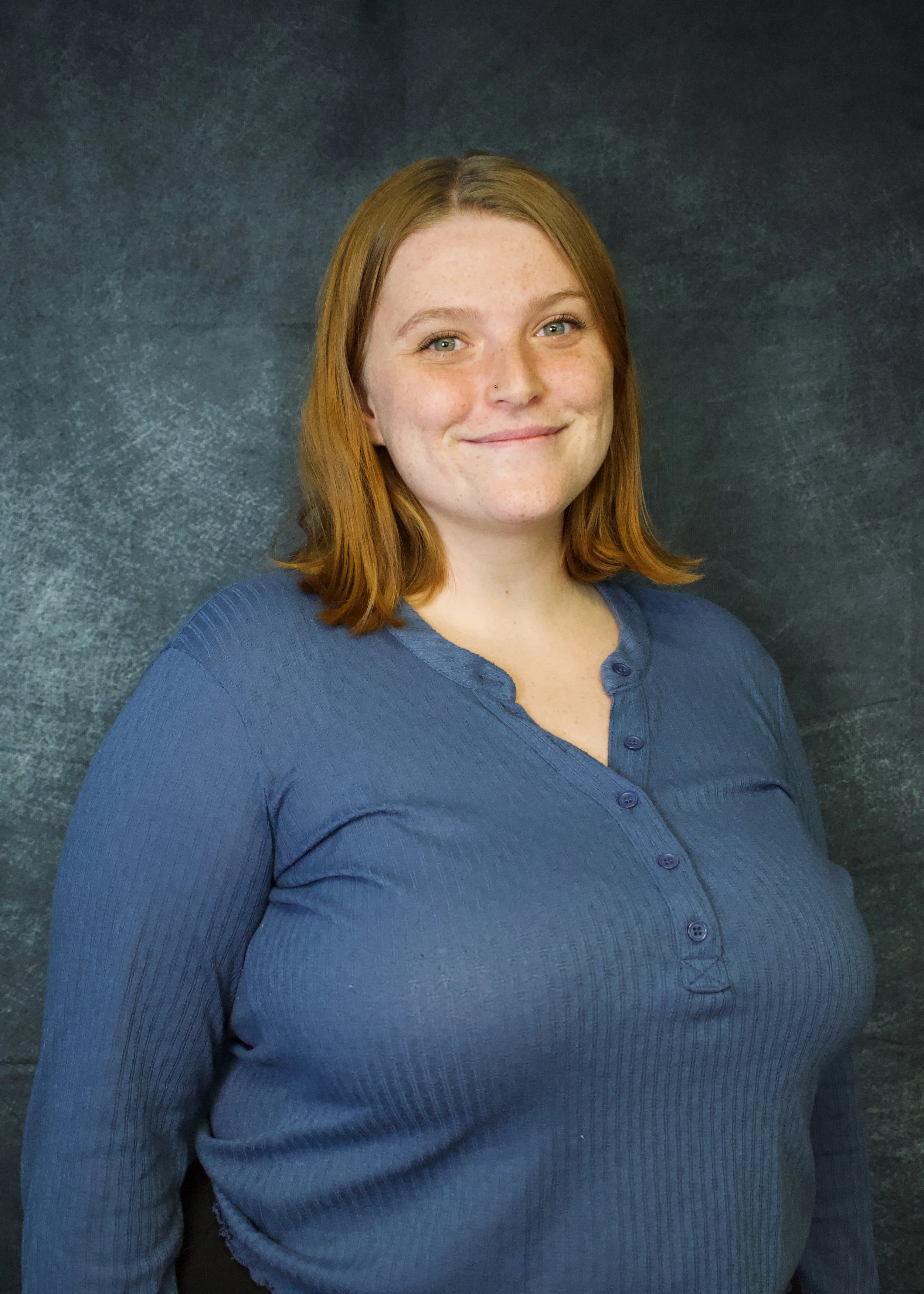 A woman with shoulder-length red hair and fair skin stands in front of a dark textured background, smiling subtly and wearing a long-sleeved blue shirt.