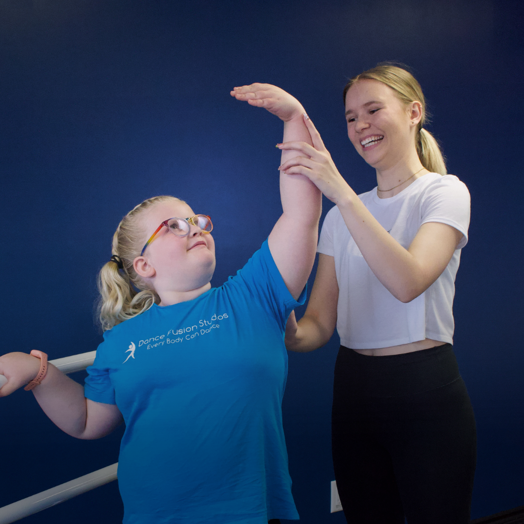 A young girl with glasses is receiving a stretch from an instructor in a dance studio with a dark blue wall.