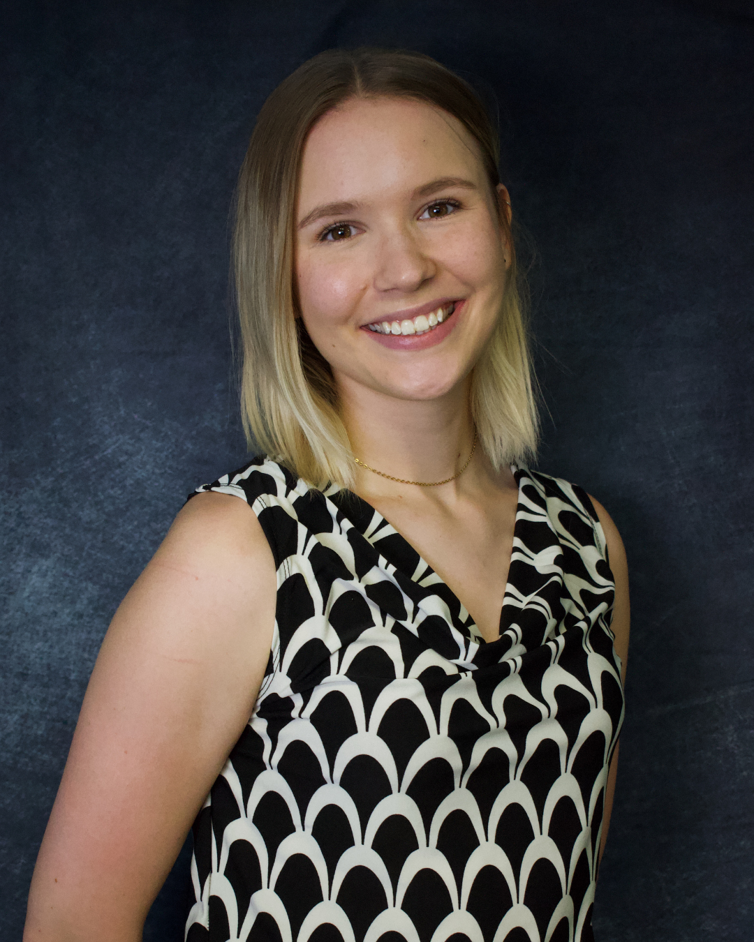 A woman with shoulder-length blonde hair and fair skin smiling, wearing a black and white patterned sleeveless top and a delicate gold necklace, standing against a dark textured background.