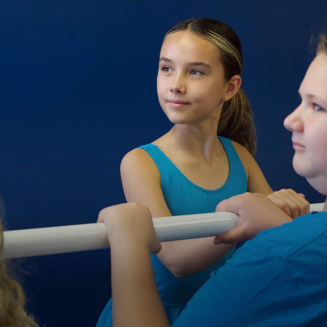 A young girl in a blue dress watching gymnast training at a ballet barre.
