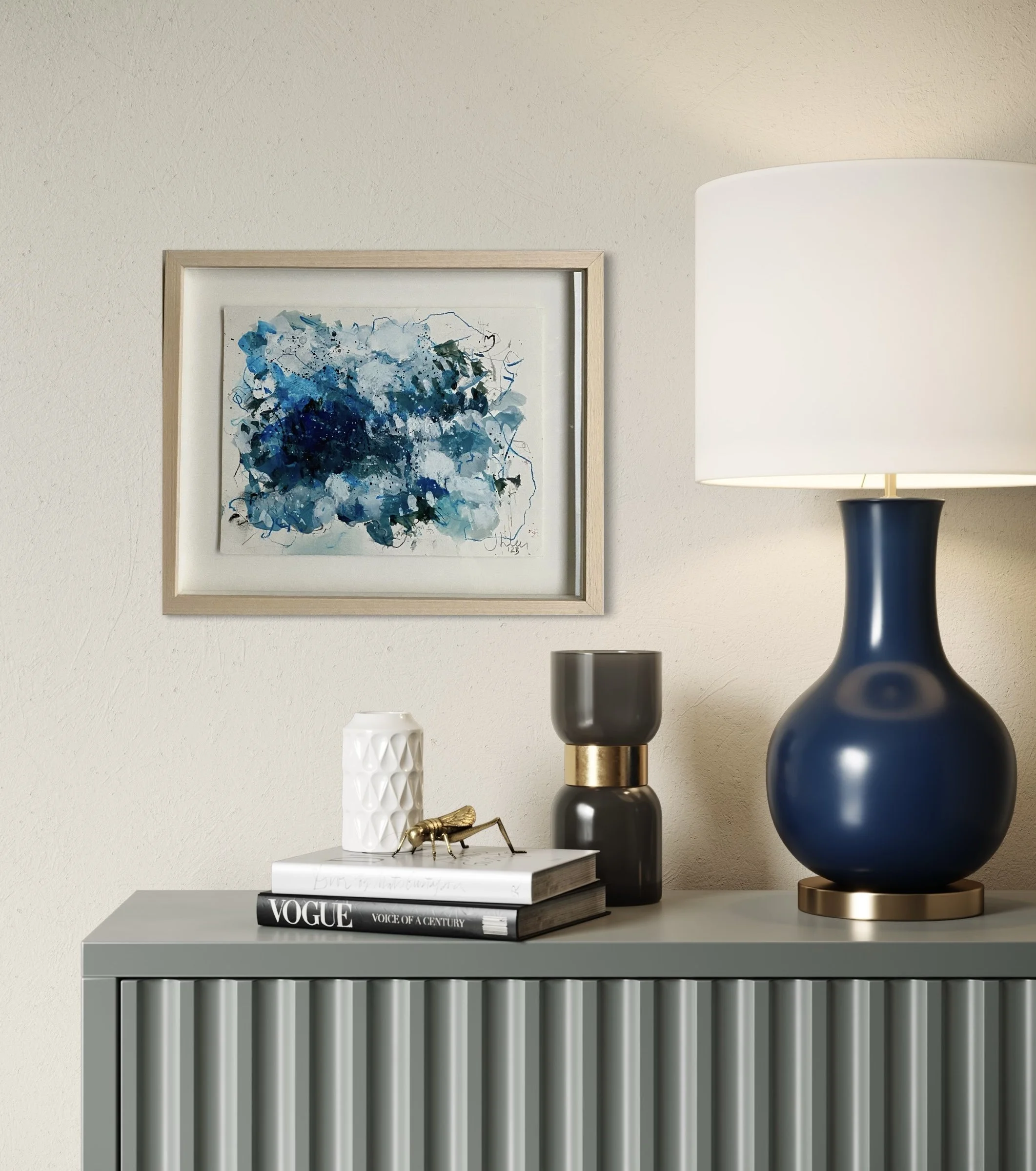 A modern living room or hallway corner with a decorative console table displaying vases, a magazine, and a decorative insect figurine. A framed abstract blue artwork hangs on the beige wall, and a large blue and white ceramic table lamp is on the right.
