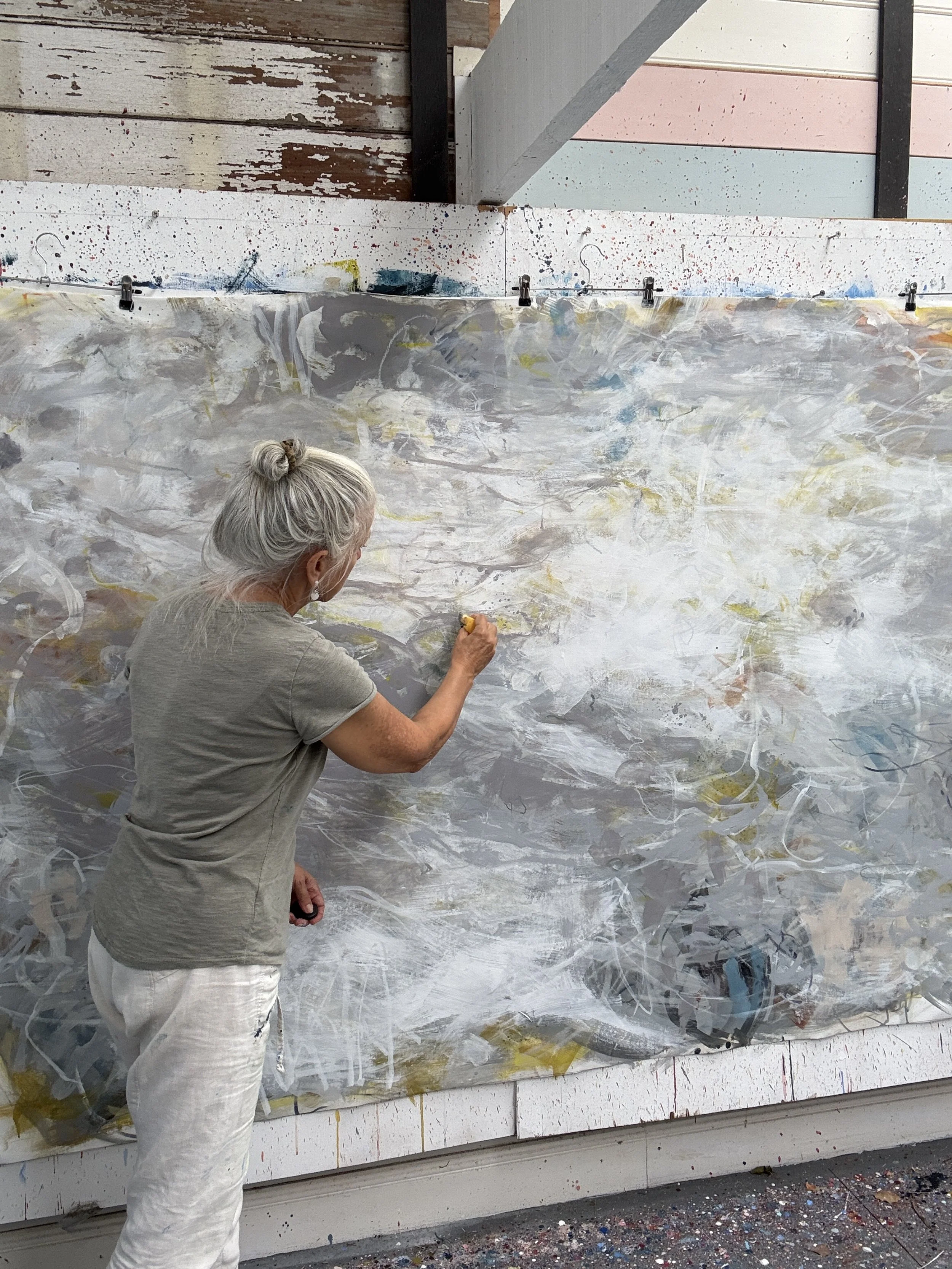 An elderly woman with gray hair tied in a bun is painting an abstract mural on a large canvas in an art studio, with colorful swirls and streaks of paint.