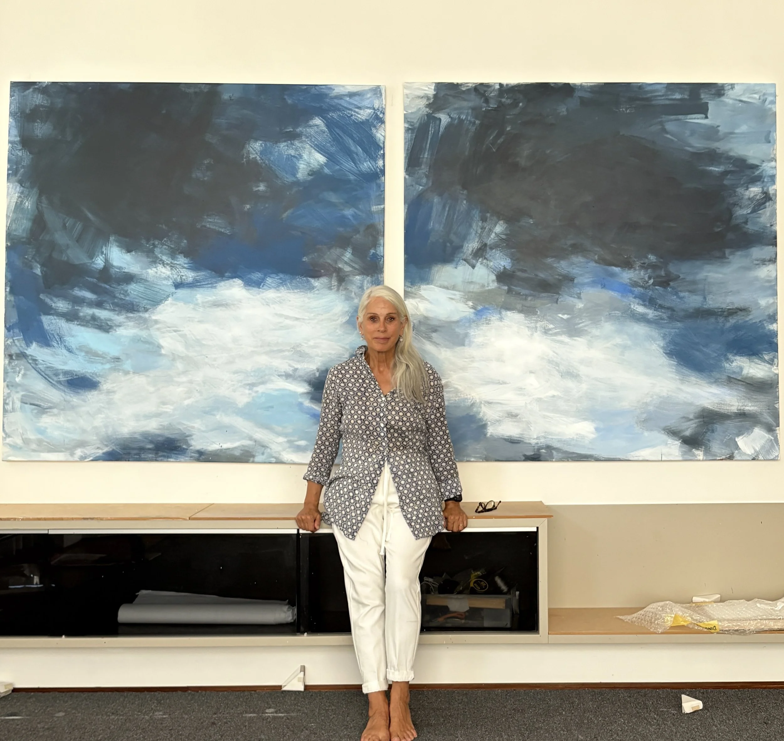 An elderly woman with long white hair, dressed in a patterned top and white pants, standing barefoot in front of two large abstract blue and white paintings, leaning on a table with some glasses and items on it.