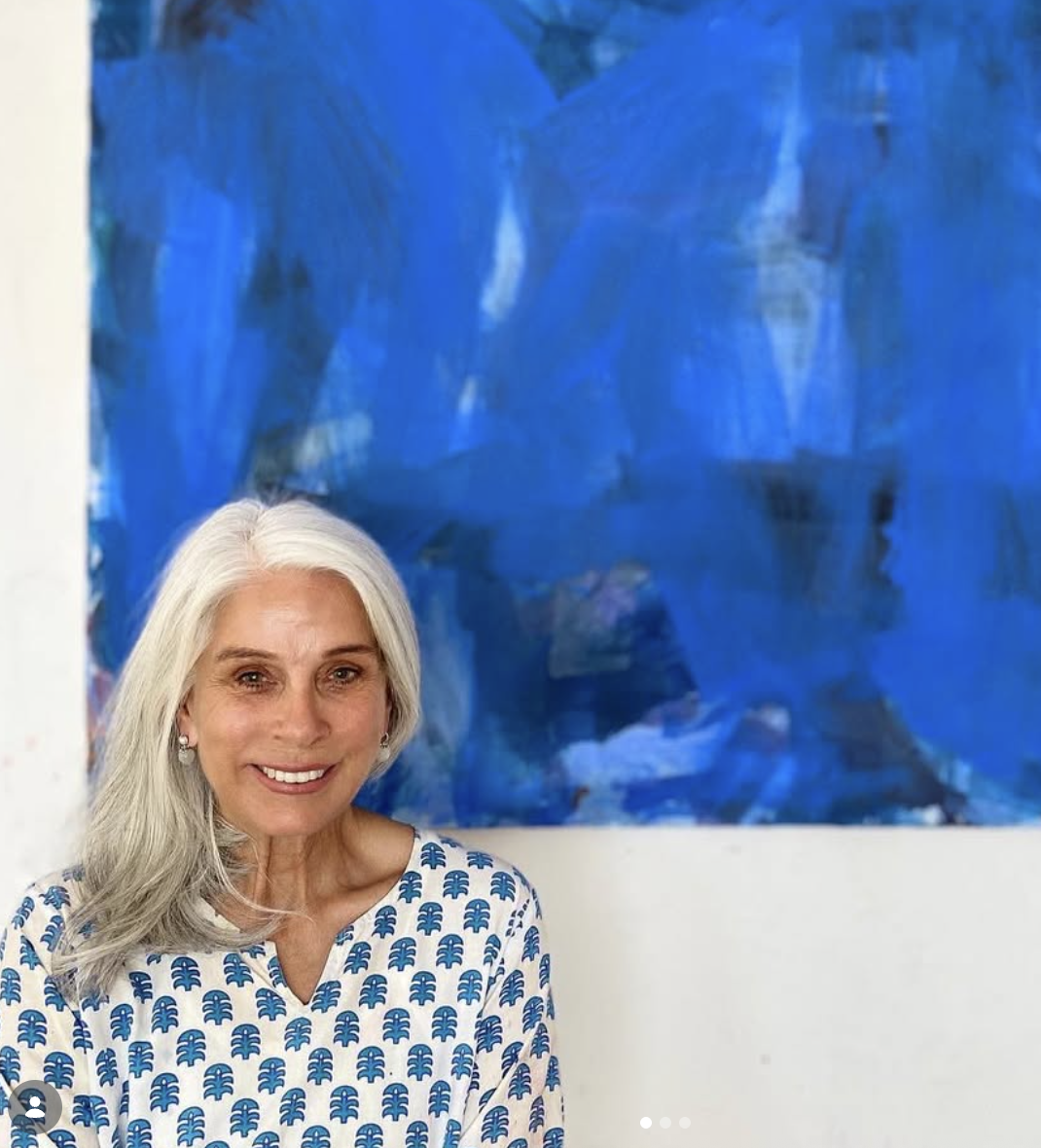 A smiling woman with long gray hair standing in front of a large abstract blue painting, wearing a white top with blue leaf patterns.